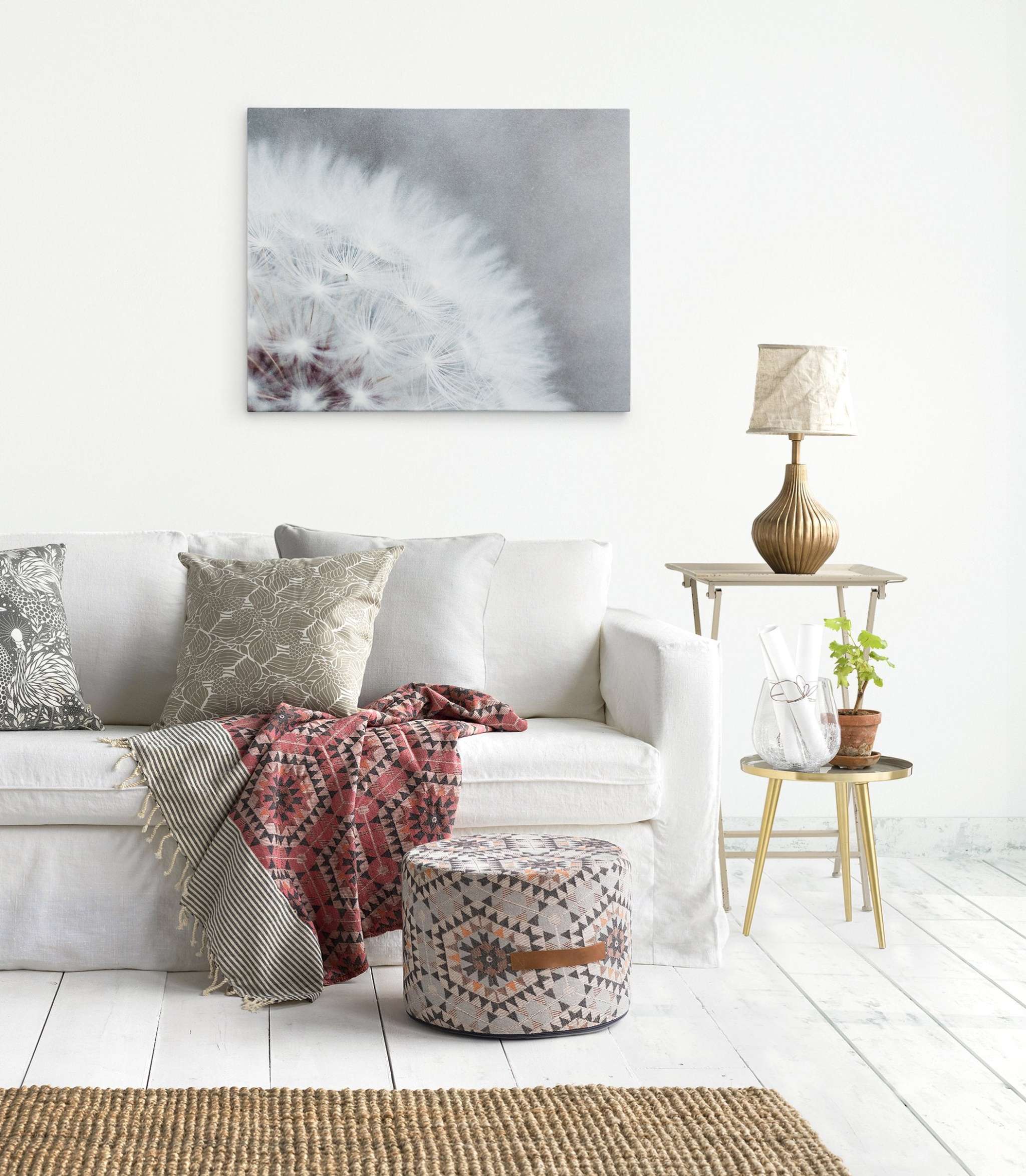 A light and airy living room features a white sofa adorned with patterned throw pillows and a red and orange blanket. Next to the sofa is a small side table with a lamp, a plant, and a glass jug. A pouf sits on the wooden floor, and above, an exquisite dandelion art piece on premium artist-grade canvas hangs on the white wall: the Grey Botanical Canvas Wall Art, 'Dandelion Queen' by Offley Green.