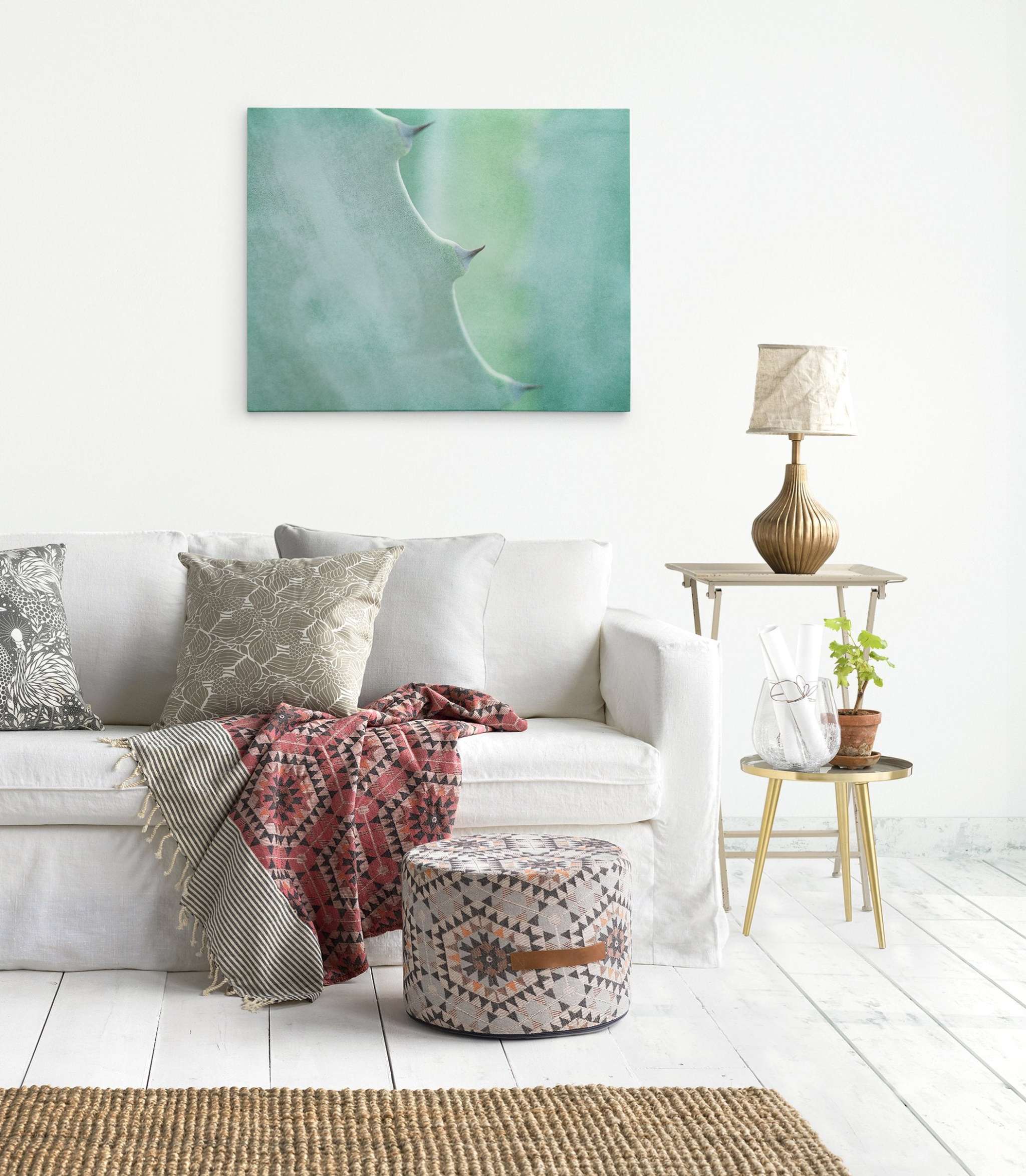 A bright and airy living room features a white sofa adorned with patterned cushions and a red geometric throw. A stylish pouf sits nearby. A side table holds a lamp, a glass pitcher, and an Aloe Vera plant. Offley Green&#39;s Mint Green Botanical Wall Art, &#39;Aloe Vera Spikes&#39; on premium artist-grade canvas hangs on the wall. Light wooden flooring.