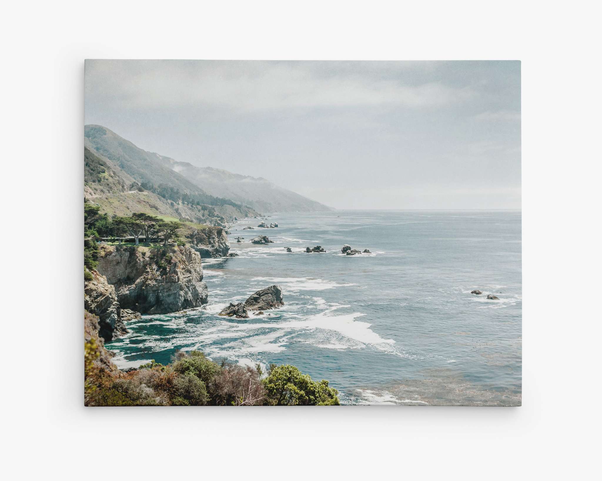 A scenic coastal view with rocky cliffs extending into the ocean under a partly cloudy sky. Waves crash against the shore, and lush green vegetation is visible in the foreground, giving way to mountainous terrain in the distance. This breathtaking coastal scene is perfect for Big Sur Landscape Canvas Wall Art, 'Rocky Rocks' by Offley Green.