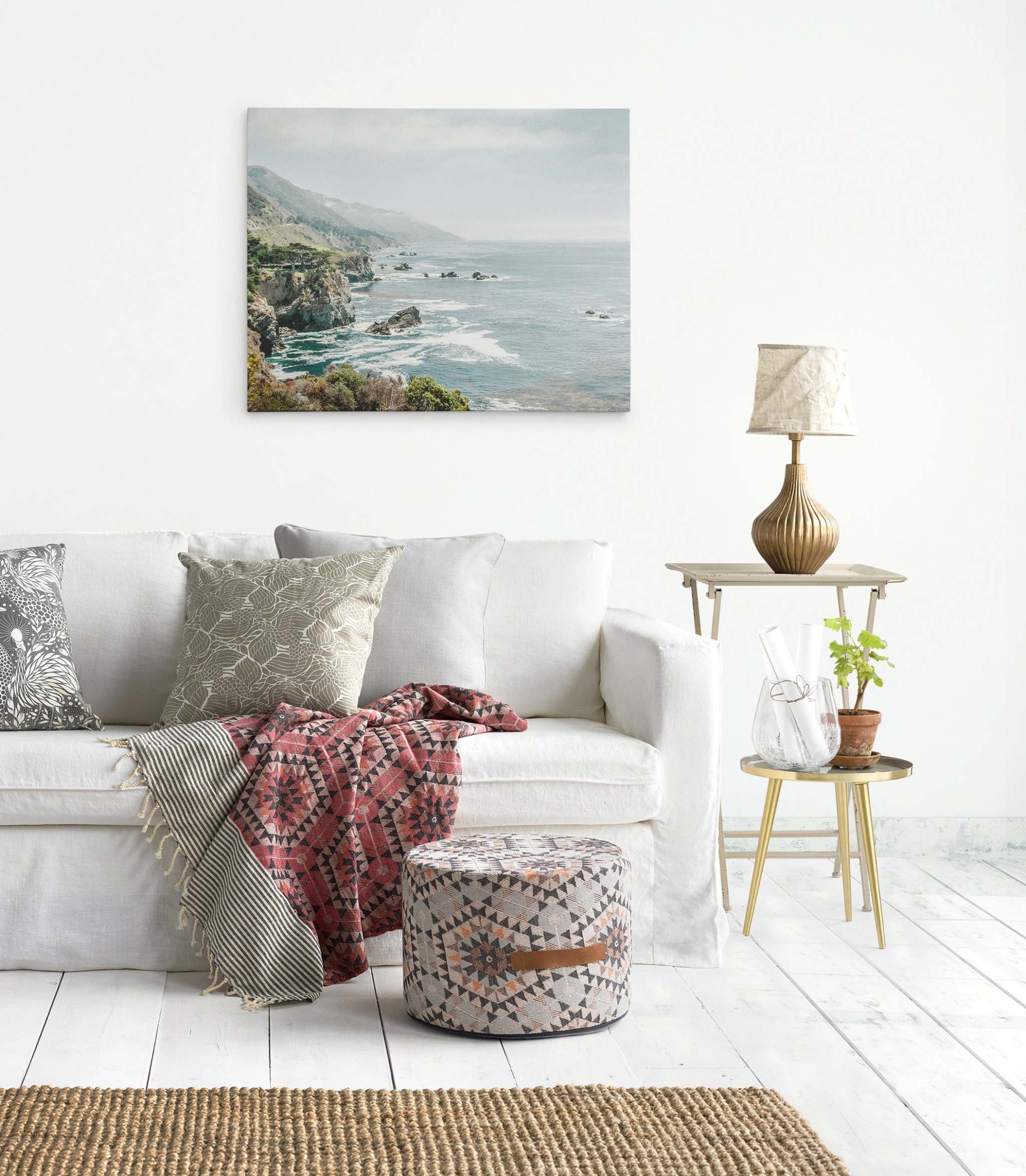 A cozy living room with a white sofa adorned with patterned pillows, a red and pink patterned blanket, and a matching pouf. A side table holds a lamp and a potted plant. A serene Big Sur Landscape Canvas Wall Art, 'Rocky Rocks' by Offley Green hangs on the wall above the sofa. The floor is white wood.