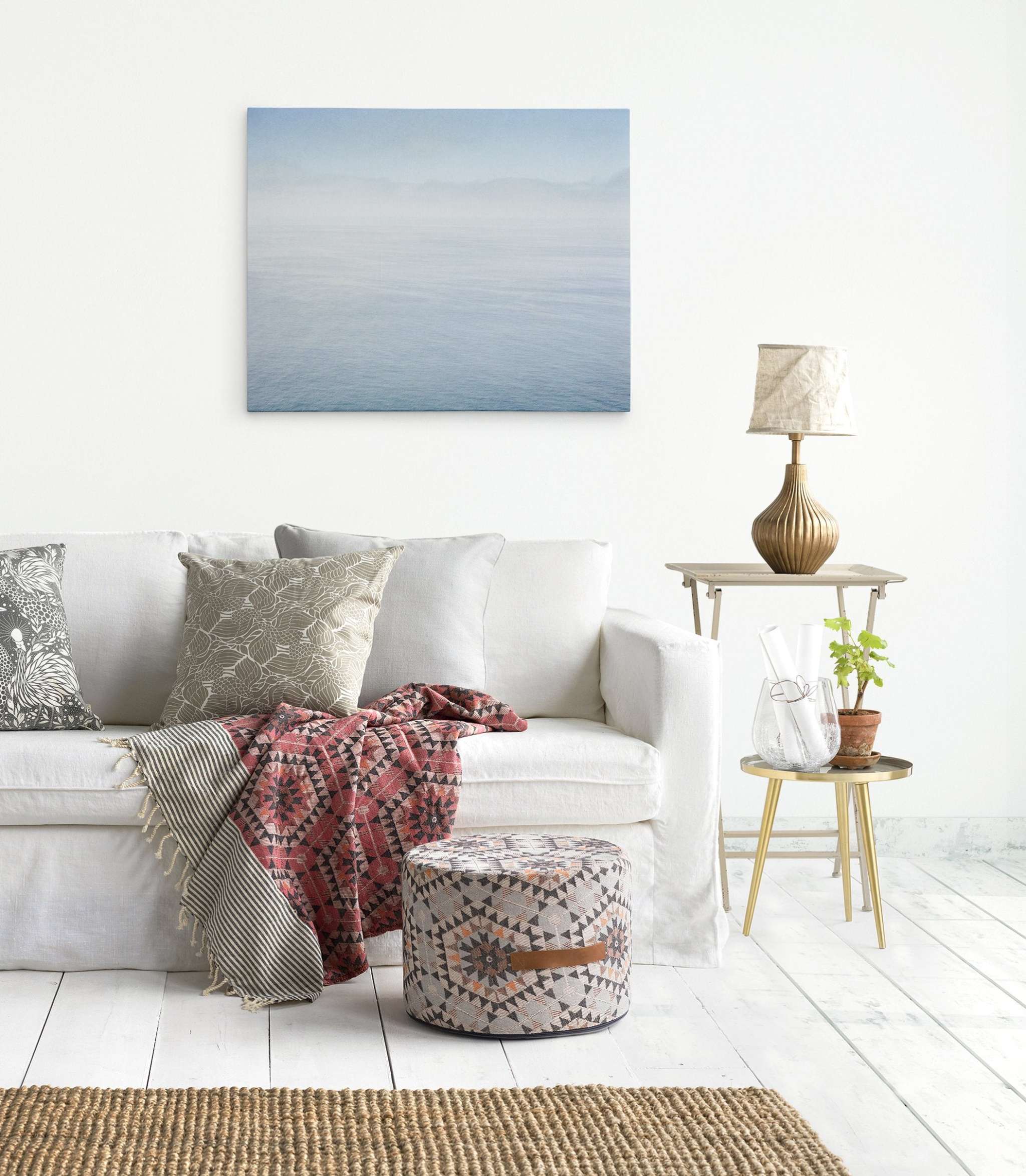 A cozy living room with a white sofa adorned with patterned pillows and a red throw. A textured ottoman sits in front, and a small side table with a lamp, plant, and glass jars is nearby. An abstract wall art of the Big Sur ocean hangs on the white wall, adding to the serene ambiance—Abstract Blue Ocean Art 'Soothing Blue Sea' by Offley Green.