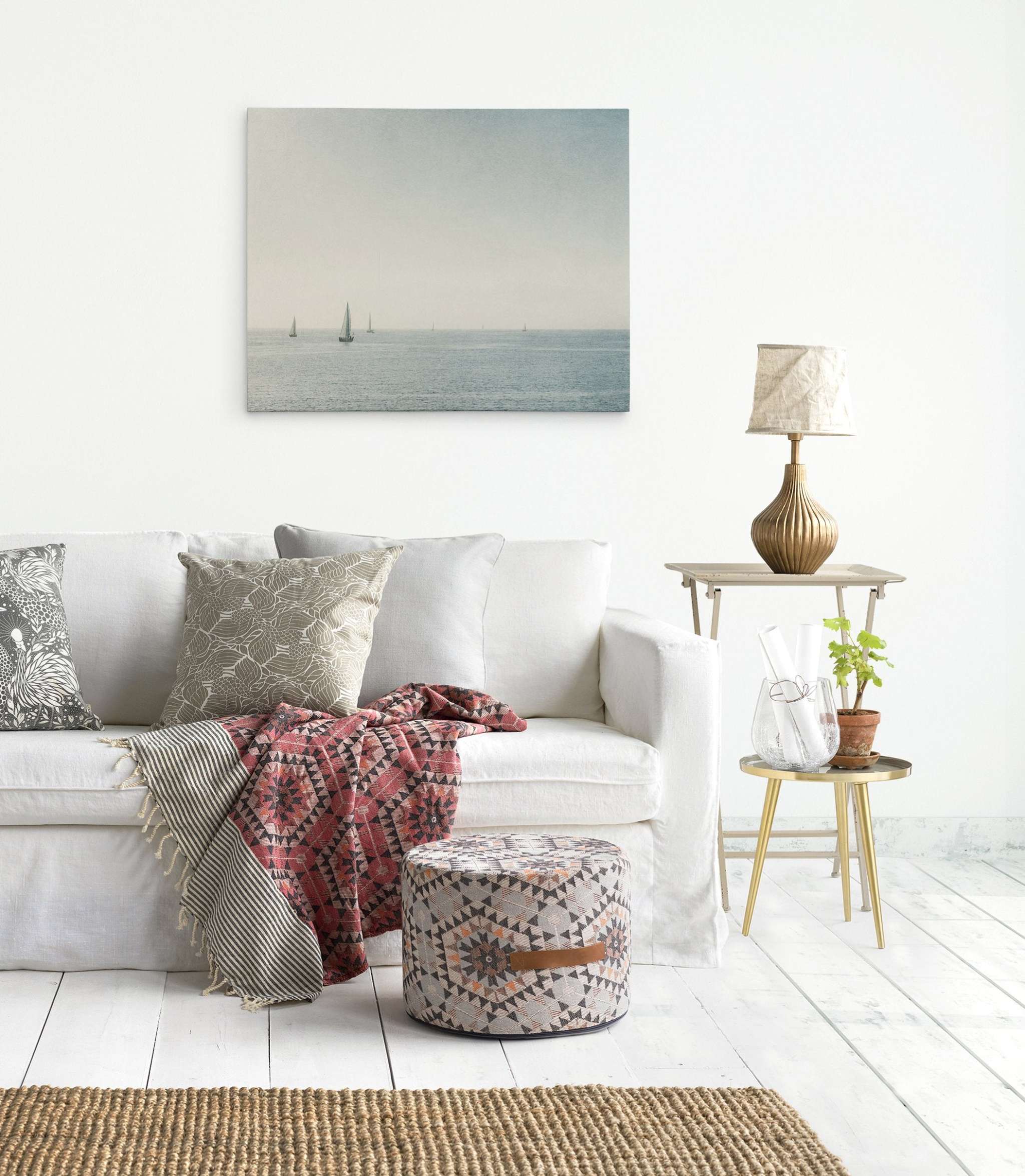 A cozy living room in Marina Del Rey features a white sofa adorned with patterned cushions and a red blanket. A pouf with geometric designs sits in front of it. A small side table holds a plant and a gold lamp, while an Offley Green Moody Nautical Seascape Canvas, 'Sail Boats Approaching' elegantly hangs on the white wall.