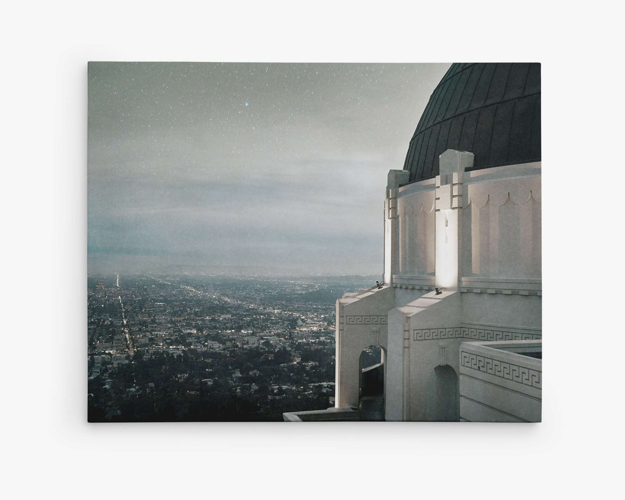 A nighttime City view of Los Angeles from the Griffith Observatory. The observatory's structure is visible on the right, partially illuminated, with the sprawling city lights stretching out below under a starry sky. The horizon is hazy, blending into the night. Perfect for Offley Green's Los Angeles Griffith Observatory Canvas Wall Decor, 'The Sky At Night'.
