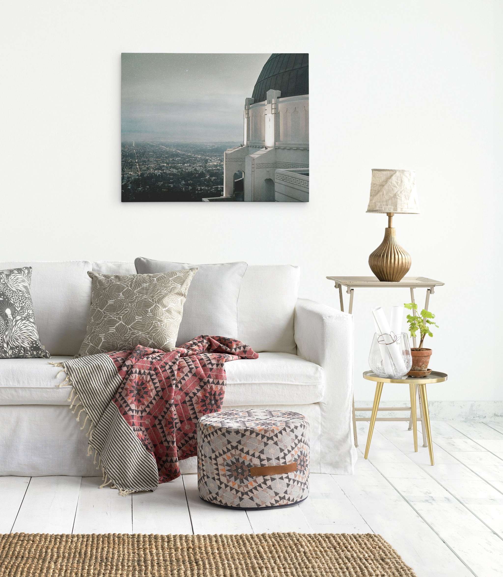 A modern living room features a white sofa adorned with patterned cushions and a red geometric throw blanket. A small gray ottoman and a side table with a gold lamp, glass vase, and plant are nearby. Los Angeles Griffith Observatory Canvas Wall Decor, &#39;The Sky At Night&#39; by Offley Green hangs on the white wall above the sofa.
