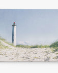 A tall, white lighthouse with a red top stands prominently against a clear blue sky. Surrounded by sand dunes and tall grasses, the serene coastal scene is perfect for capturing in a lighthouse painting. This setting would make stunning Offley Green Nautical Canvas Wall Art, 'The Lighthouse' or a beautiful canvas gallery wrap.