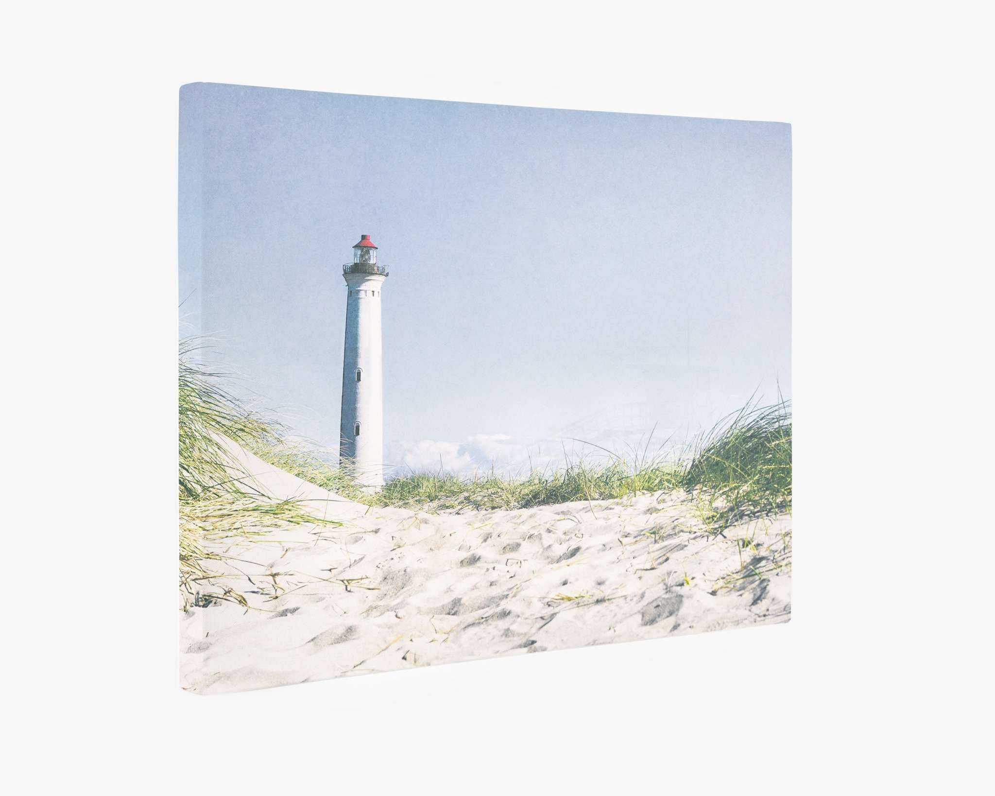 A vertical canvas gallery wrap displays a serene beach scene with a tall white lighthouse in the distance. Sand dunes and beach grass are in the foreground under a clear, pale blue sky, making it Offley Green&#39;s Nautical Canvas Wall Art, &#39;The Lighthouse.&#39;