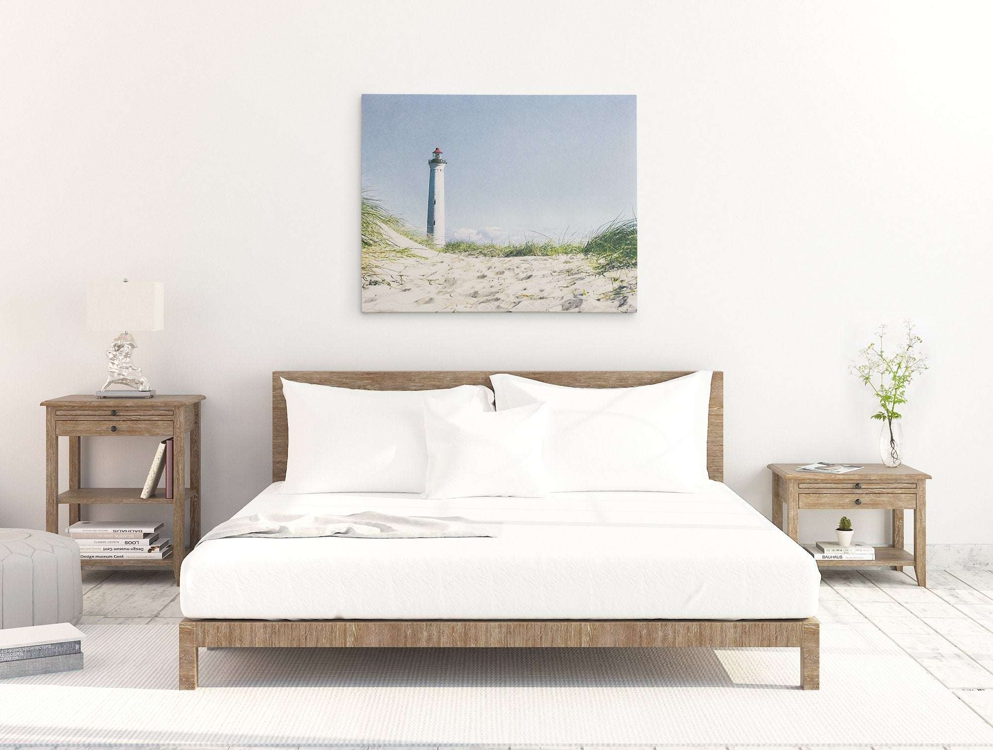 A minimalist bedroom with a wooden bed frame and white bedding. Two wooden nightstands flank the bed, each with a lamp and various decor. Above the bed, a large **Offley Green Nautical Canvas Wall Art, &#39;The Lighthouse&#39;** is displayed. The room has a bright, airy feel with white walls and a light floor.