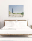 A minimalist bedroom with a wooden bed frame and white bedding. Two wooden nightstands flank the bed, each with a lamp and various decor. Above the bed, a large **Offley Green Nautical Canvas Wall Art, 'The Lighthouse'** is displayed. The room has a bright, airy feel with white walls and a light floor.