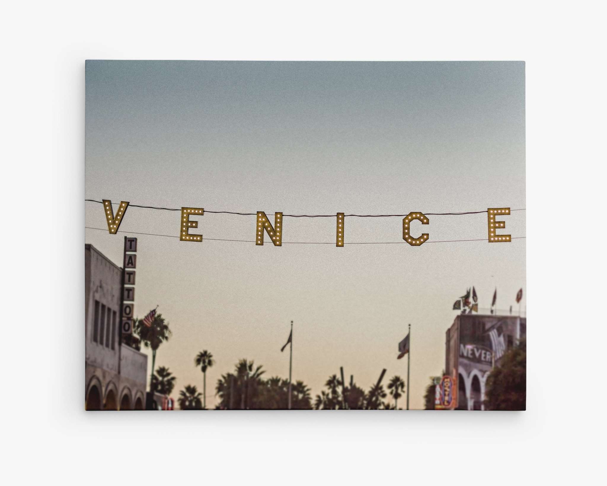 A sunset sky frames the illuminated Venice Beach sign strung across a street. Palm trees line the background, and partial views of buildings, including a tattoo shop and other storefronts, are visible, adding to the coastal city's iconic atmosphere, perfect for the California Venice Beach Sign Canvas Wall Art, 'Venice Sunset' by Offley Green.
