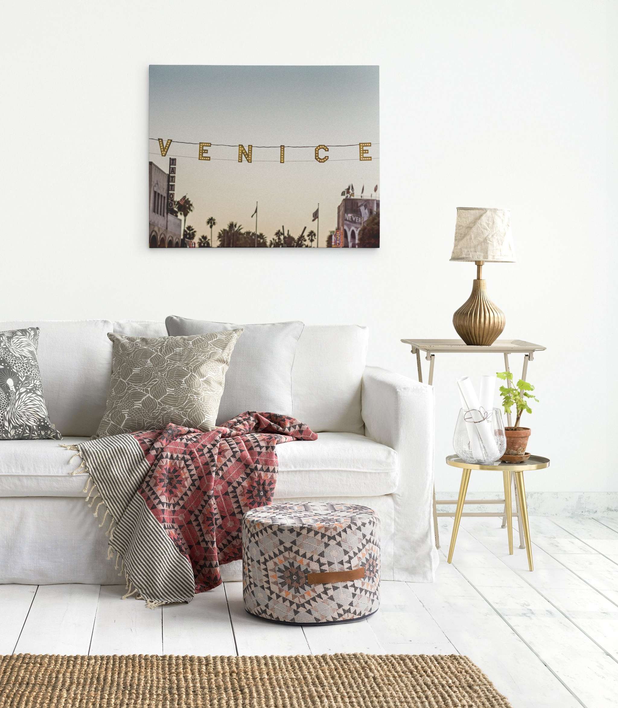 A cozy, stylish living room features a white sofa with patterned cushions and a throw blanket, a round patterned ottoman, and a side table with a lamp and plant. A premium Offley Green artist-grade canvas above the sofa reads "California Venice Beach Sign Canvas Wall Art, 'Venice Sunset'." The light wood floor and woven rug complete the inviting space.