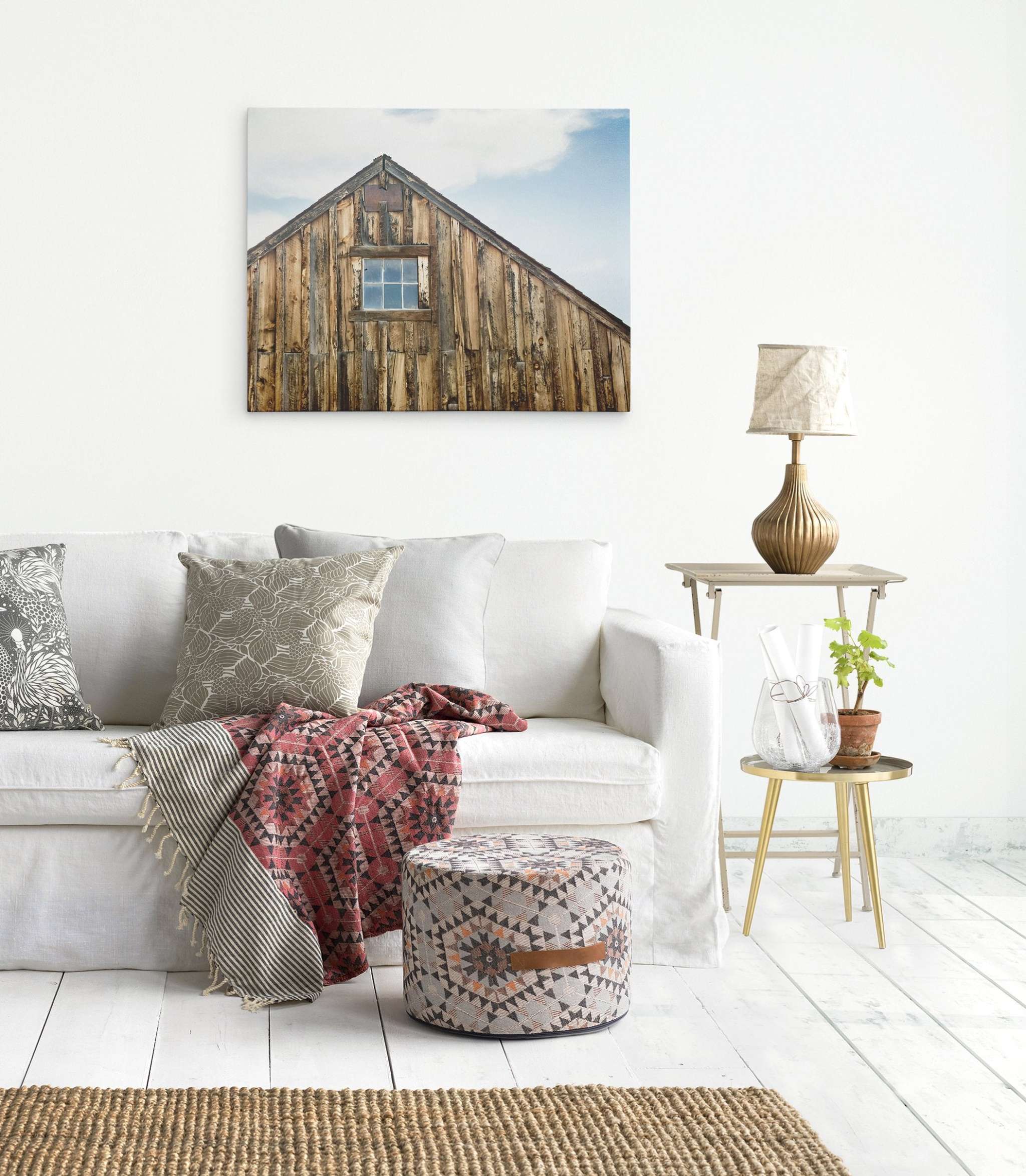 A cozy living room with a white sofa adorned with various patterned pillows and a red patterned throw blanket. In front of the sofa is a patterned ottoman. To the right, a small wooden side table holds a lamp and potted plant. Farmhouse Wall Art, Rustic Barn Decor, 'Old Barn at Bodie' by Offley Green hangs above the sofa.