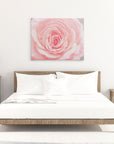 A minimalist bedroom with a wooden bed frame, white bedding, and a large Offley Green Pink Rose Canvas Wall Art, 'Pink and Shabby' on the wall. There are two matching wooden nightstands on either side, each with a lamp and decorative items. The room has a bright and airy feel.