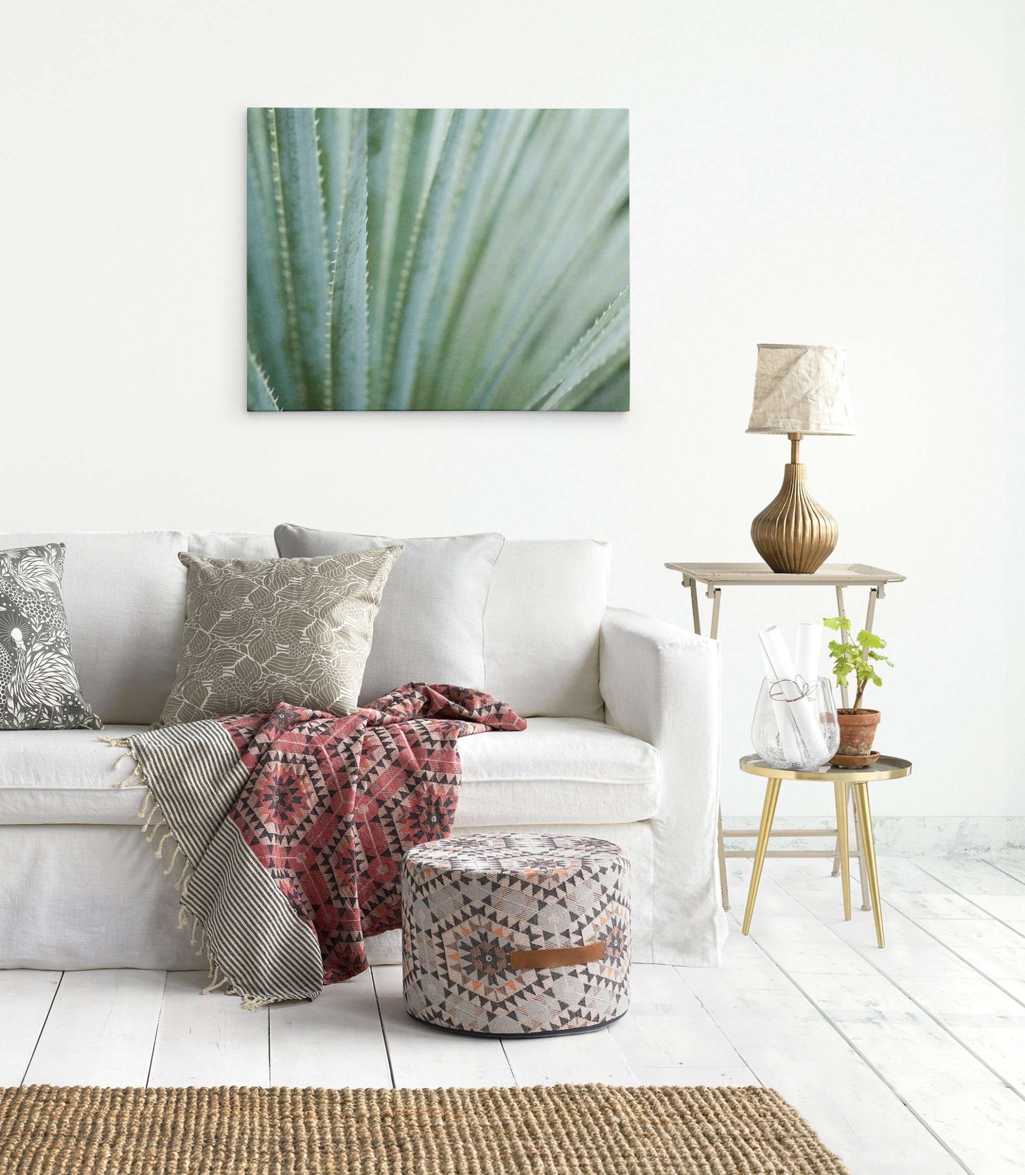 A cozy living room corner with a white sofa adorned with various patterned pillows and a red throw, a small wooden side table with a lamp, and an Offley Green &#39;Strands and Spikes&#39; Abstract Green Botanical Canvas Wall Art on the wall.