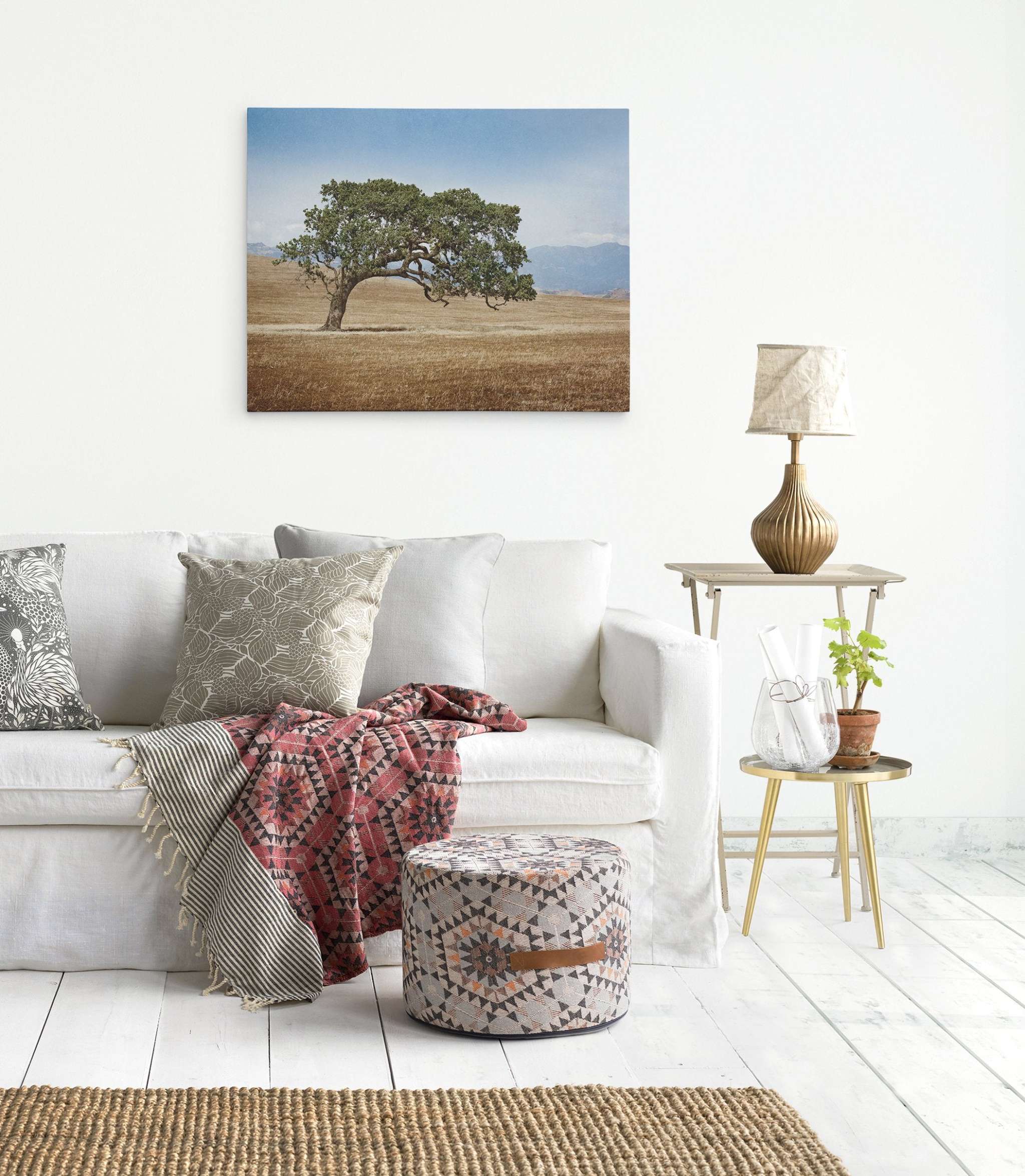 A cozy living room with a white couch adorned with patterned pillows and a colorful throw blanket. A patterned ottoman is placed in front. A side table with a plant, vase, and lamp is next to the couch. A large framed Offley Green California Landscape Wall Art, Oak Tree Canvas 'Windswept' hangs on the wall.