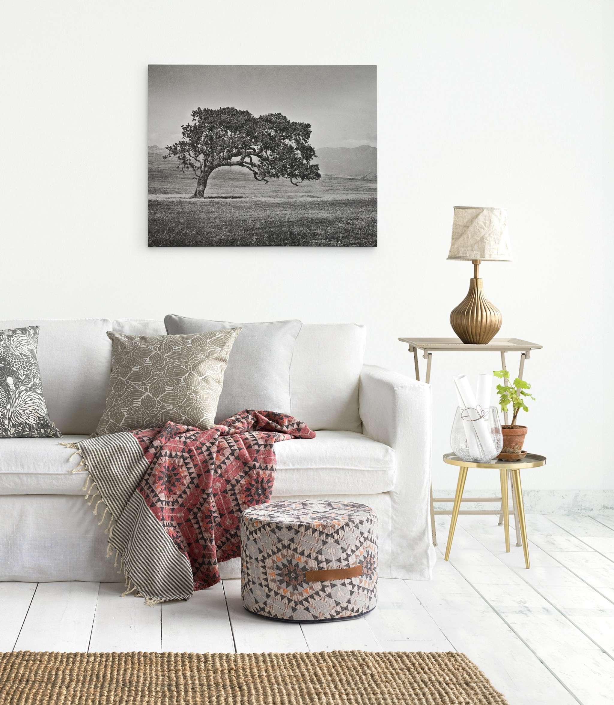 A bright living room features a white sofa with patterned and plain cushions, and a decorative blanket. A patterned ottoman sits in front. Beside the sofa, a small table holds a lamp and small potted plants. A ready-to-hang Californian Oak Tree Landscape Canvas, 'Windswept (Black and White)' by Offley Green adorns the wall.