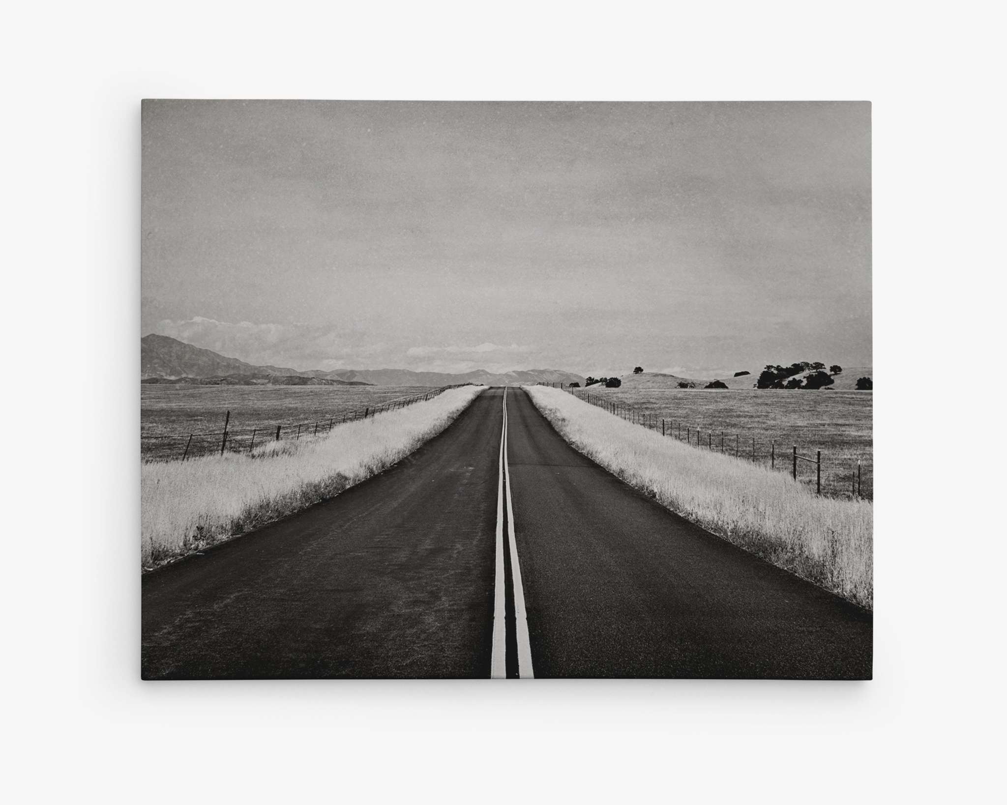 A black and white photograph of an empty, straight road stretching into the horizon. The road is flanked by fields of tall grass and sparse trees. Mountains are faintly visible in the distance under a cloudy sky, creating a serene and open landscape—perfect wall art for any space on premium artist-grade canvas. This is Offley Green's Black and White Rural Landscape Canvas Art, 'American Road Trip.'