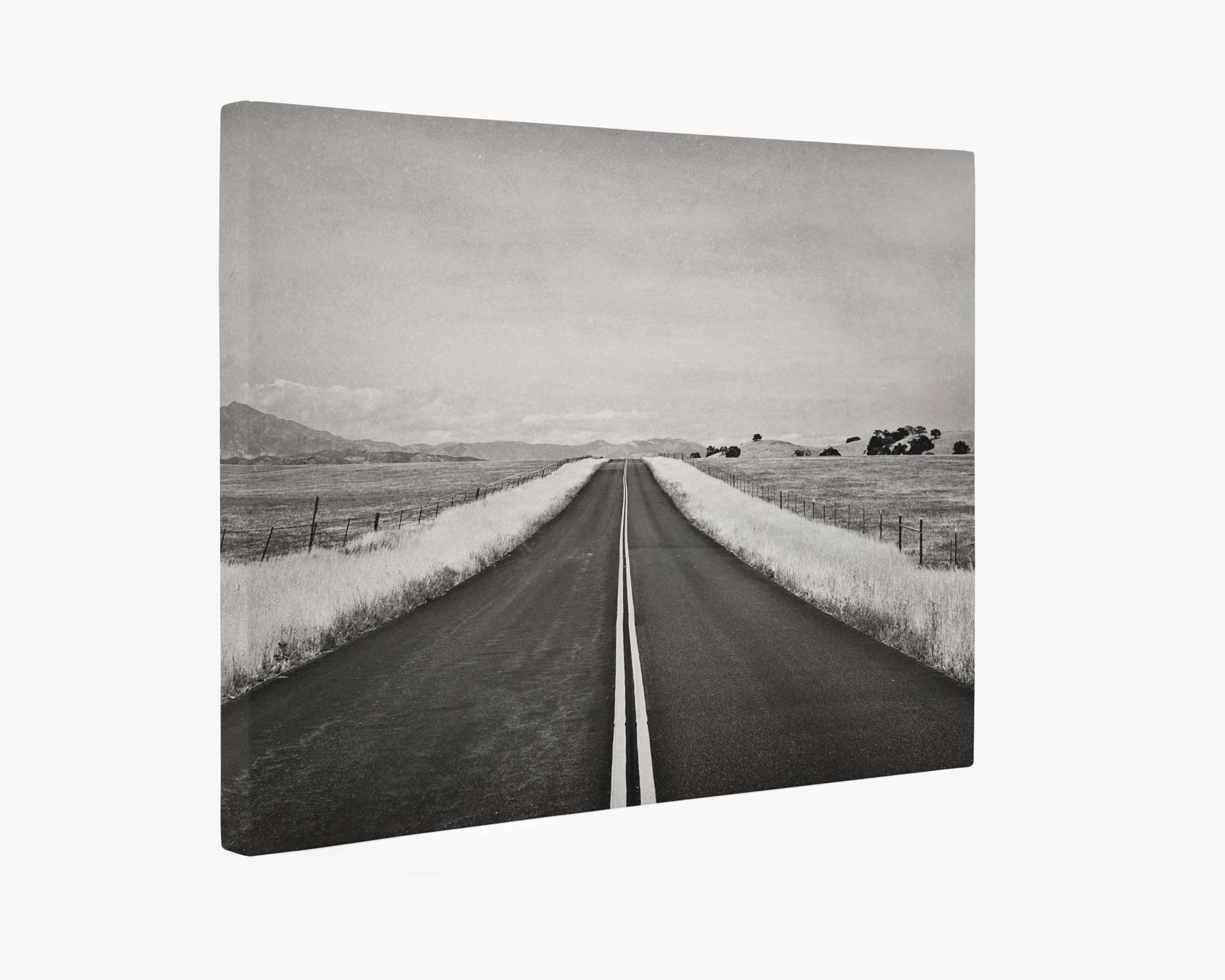 A black and white photograph, perfect as wall art, shows a long, straight road stretching into the distance. The road is flanked by grassy fields and wire fences, with distant hills barely visible under an expansive sky. This premium artist-grade canvas image, Offley Green’s Black and White Rural Landscape Canvas Art, 'American Road Trip,' evokes a sense of solitude and endless journey.
