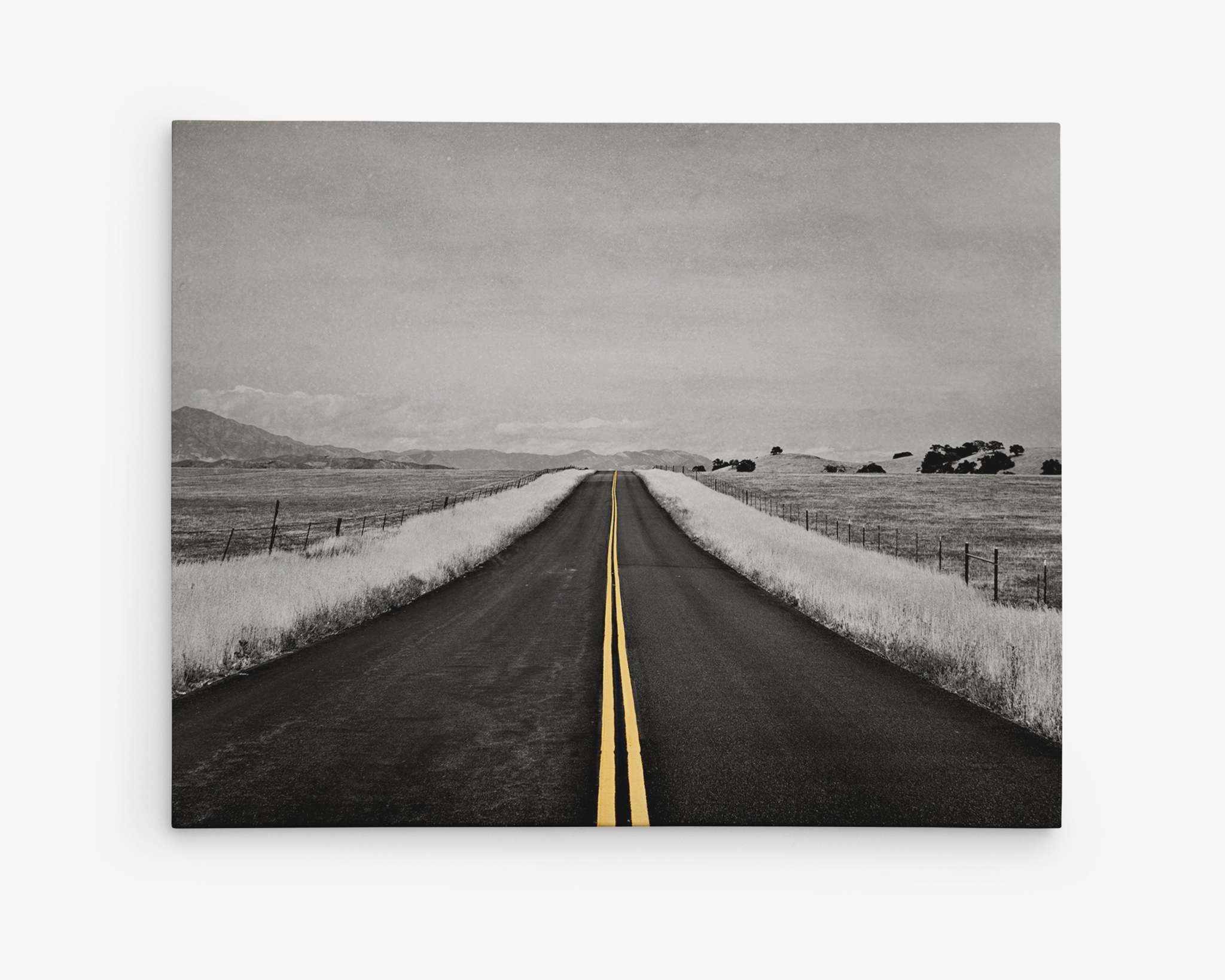 A black-and-white photograph captures a long, straight road stretching into the distance, reminiscent of a yellow brick road. The road is flanked by grassy fields and features a solid yellow line down the center. The horizon is faintly visible under a cloudy sky—ideal for striking canvas prints or wall art like Offley Green's 'Yellow Road Trip,' Black and White Road Photography with Color Accent.
