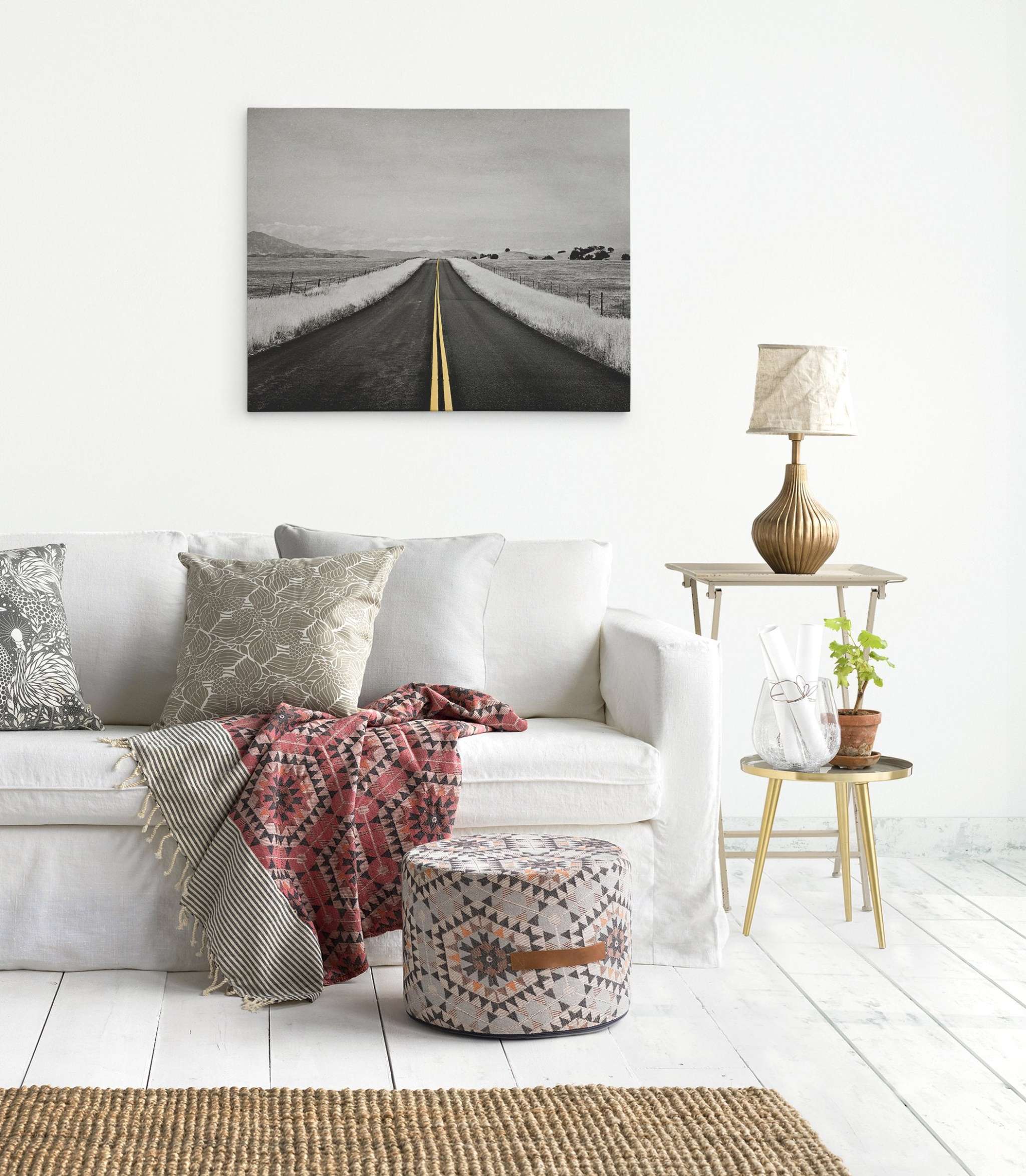A minimalist living room with a white sofa adorned with patterned cushions and a red geometric throw, a pouf with a similar design, a small side table with a lamp and plants, and large Offley Green canvas prints of Black and White Road Photography with Color Accent, 'Yellow Road Trip' on the white wall.