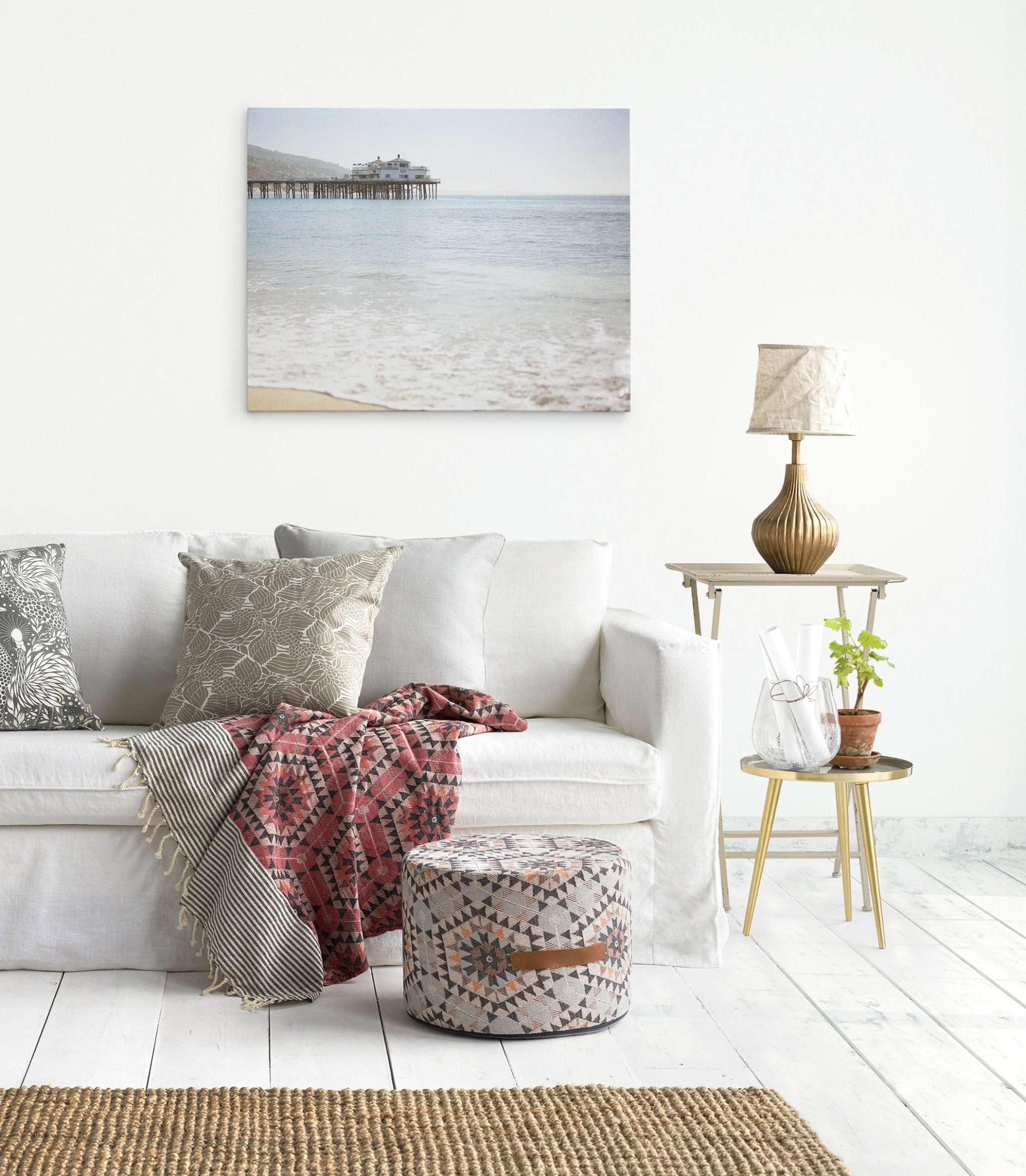 A cozy living room featuring a white sofa with assorted cushions, a patterned ottoman, wooden floor lamp, side table with a plant, and a large Offley Green Malibu Pier Wall Art on the wall.
