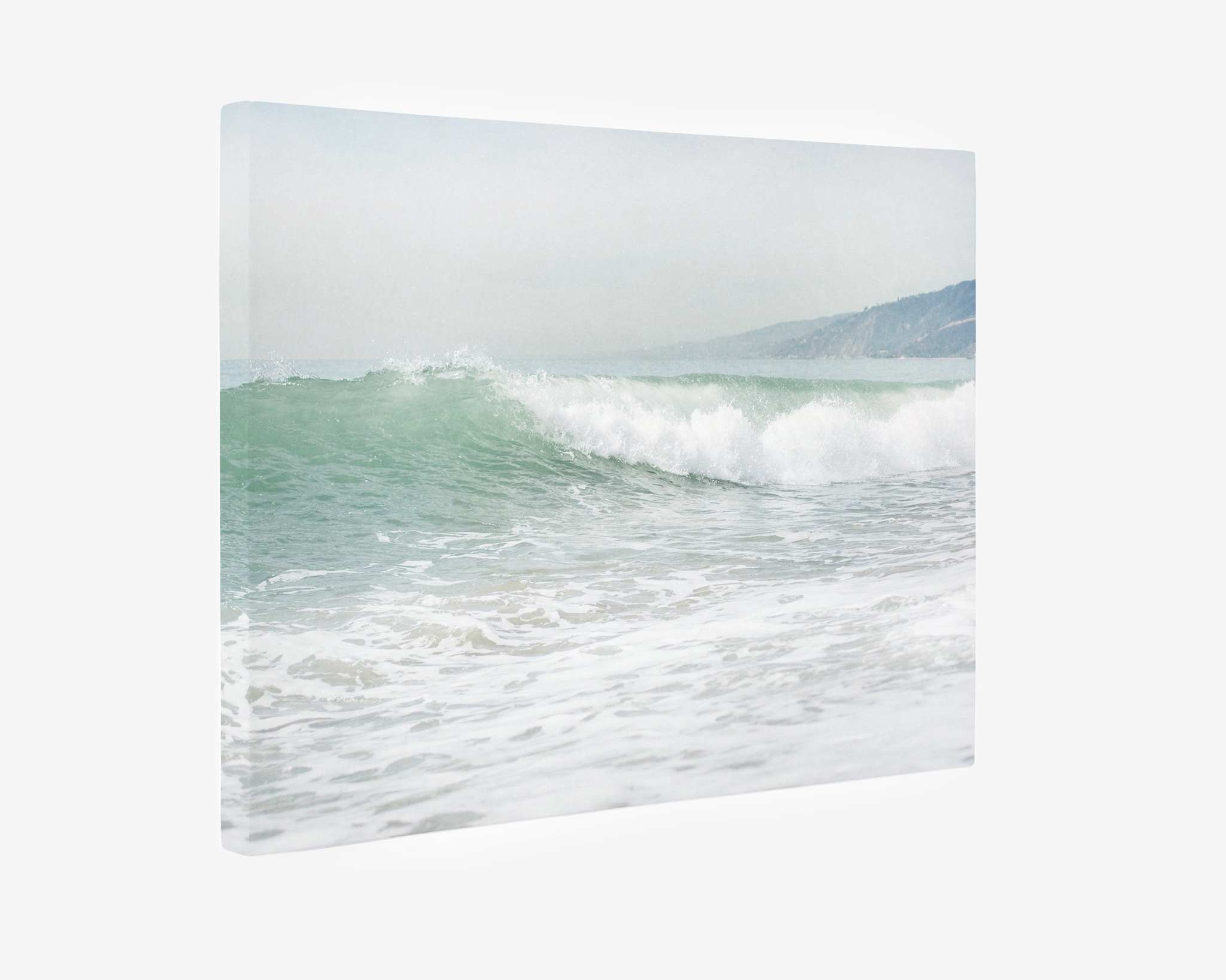 A canvas print depicts a serene coastal scene with gentle waves rolling onto the shore. The ocean water is a calm blue-green, and faint mountains can be seen in the distant background under a slightly overcast sky. This Coastal Ocean Canvas Waves Wall Art, &#39;Breaking Surf&#39; by Offley Green is crafted on premium artist-grade canvas for lasting beauty.