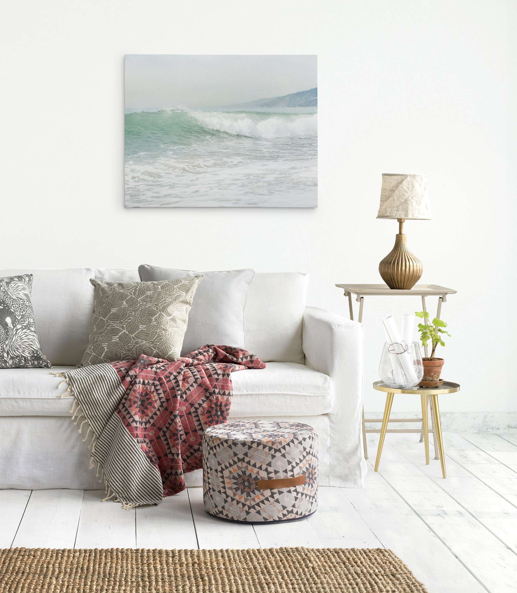 A cozy living room with a white sofa adorned with patterned throw pillows and blankets. A pouf sits in front of the sofa. A side table next to the sofa holds a rustic lamp, a plant, and decorative glassware. Beach-themed wall art on premium artist-grade canvas brings coastal sunshine to the space, like the Coastal Ocean Canvas Waves Wall Art, &#39;Breaking Surf&#39; by Offley Green.