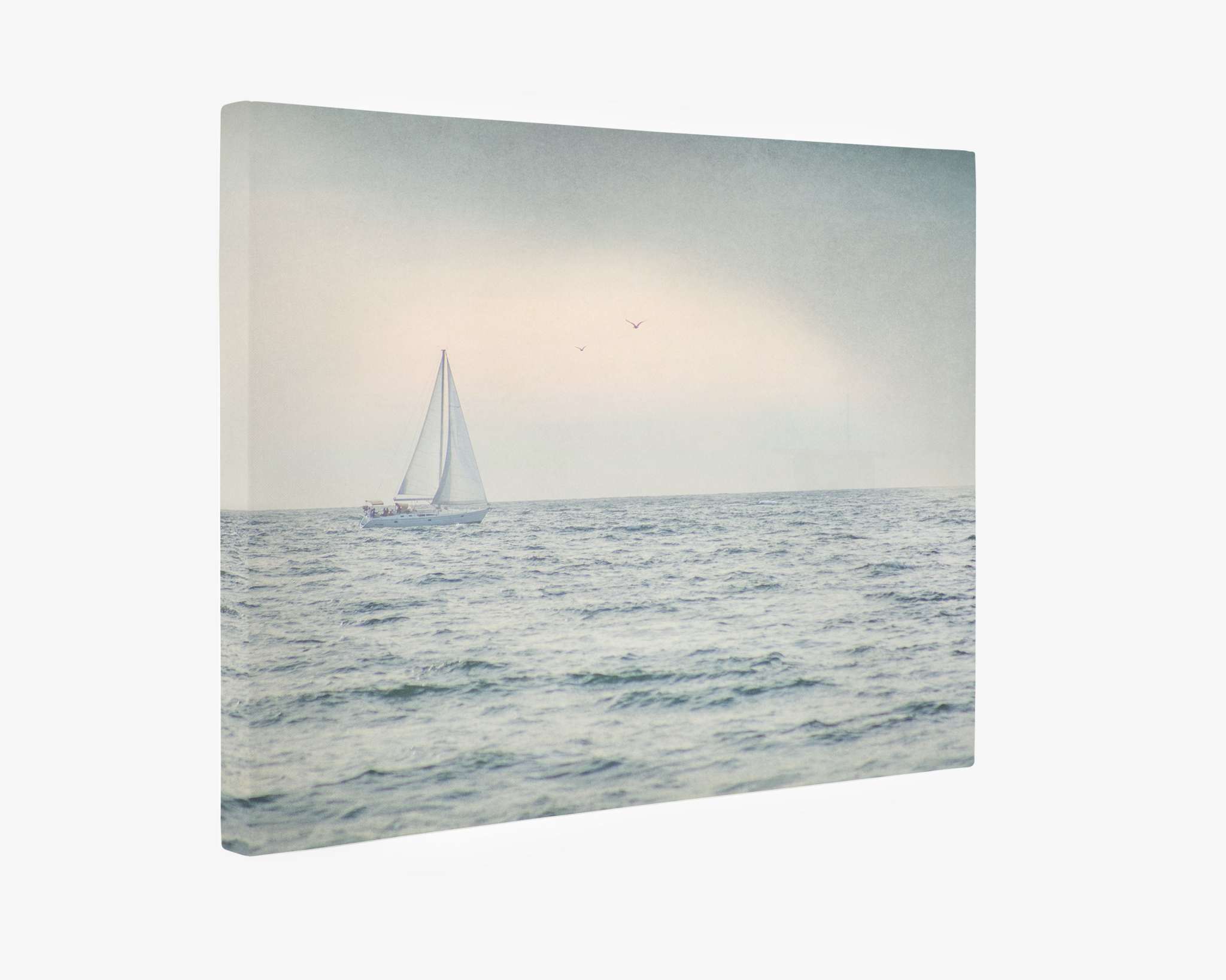 A serene seascape features a lone sailboat with white sails navigating gentle waves under a cloudy sky. Three distant seabirds fly above the horizon, adding to the tranquil scene. The Offley Green Nautical Sail Boat Canvas Art, 'Sailing Into Rain,' presented on premium artist-grade canvas, is softened with a pastel-toned filter, enhancing its calm atmosphere.