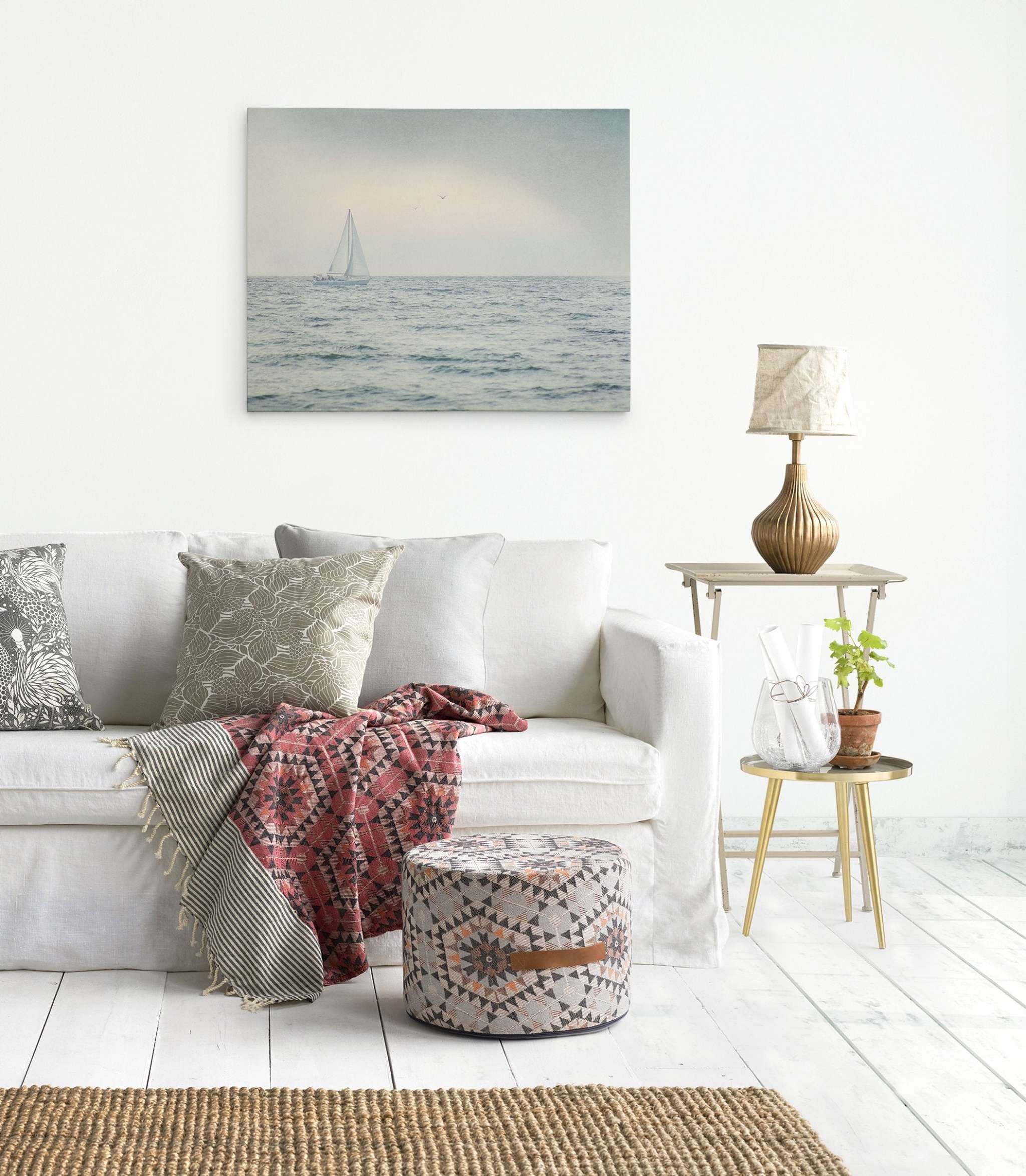 A cozy living room with a white sofa adorned with patterned pillows and a red throw blanket. A textured pouf and a side table with a lamp and plant complete the setup. Behind the sofa, Offley Green's Nautical Sail Boat Canvas Art, 'Sailing Into Rain' on premium artist-grade canvas adds an elegant touch to the white wall.