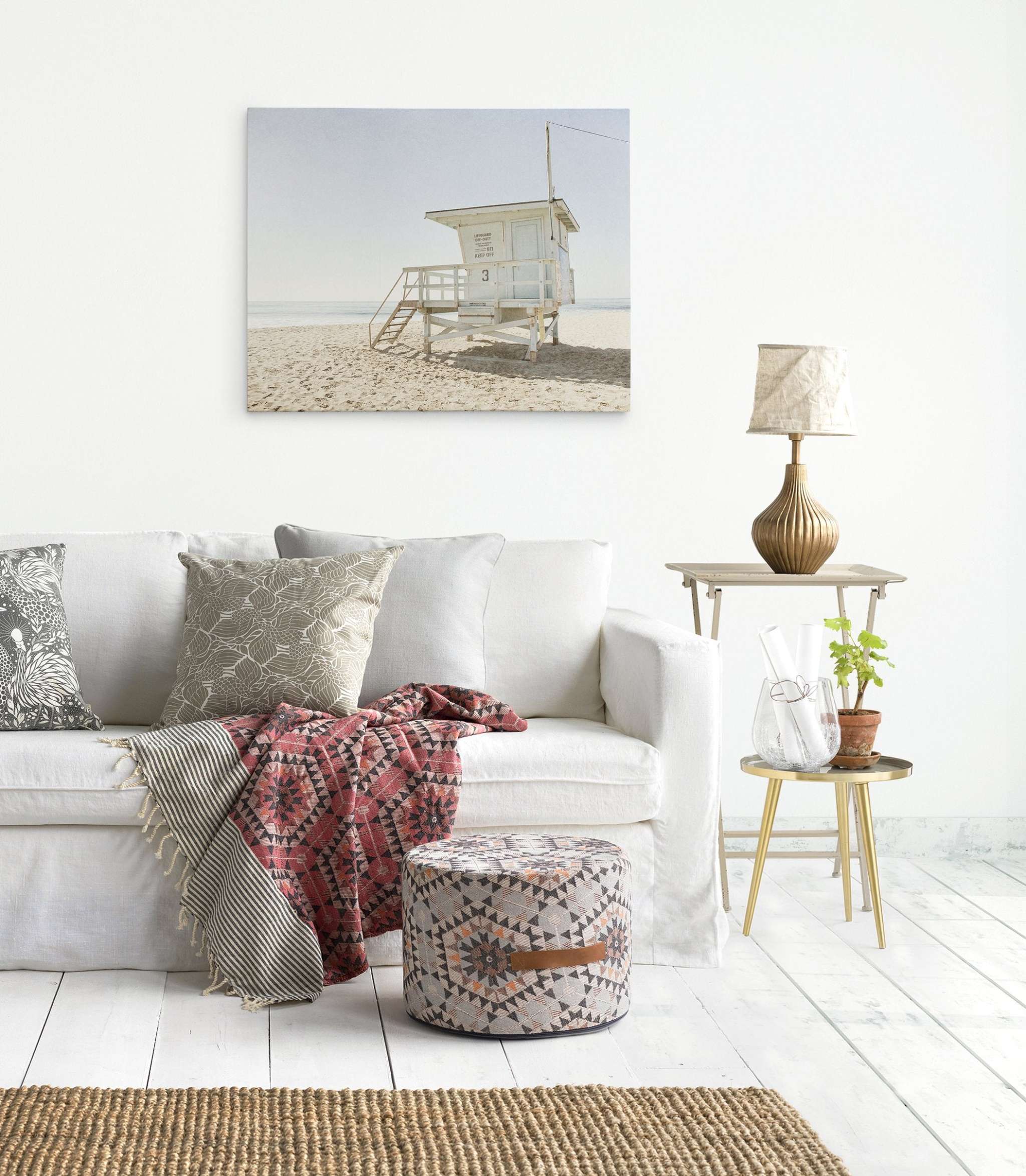 A cozy living room features a white sofa adorned with patterned pillows and a red throw blanket. In front of the sofa is a patterned ottoman. A side table holding a lamp, small plant, and glass vase stands beside the sofa. A California Summer Beach Canvas Art, 'Malibu Lifeguard Tower' by Offley Green showcases Malibu lifeguard towers under coastal sunshine California hanging on the wall.
