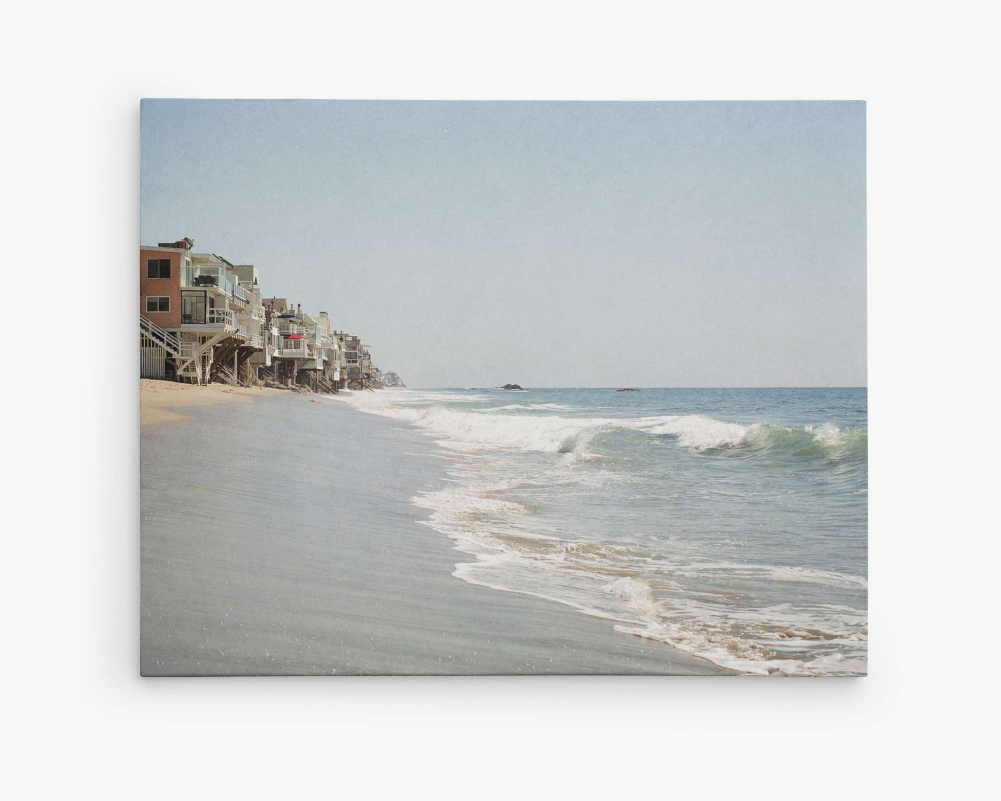 A serene beach scene with gentle waves washing up on the sandy shore. Beachfront houses on stilts line the left side, overlooking the ocean along the Malibu coastline. The sky is clear with a subtle gradient of blue, signaling a sunny day. This scene exudes tranquility and coastal charm, perfect for Offley Green's Malibu Beach House Canvas Wall Art, 'Ocean View'.