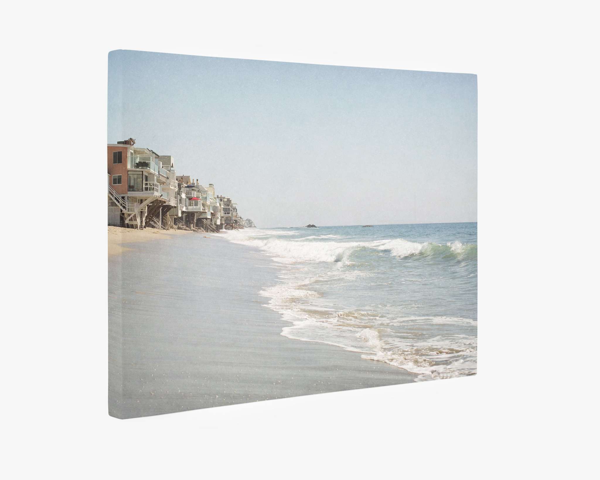 The Offley Green Malibu Beach House Canvas Wall Art, 'Ocean View' features a serene beach scene with gentle waves meeting a sandy shore. Coastal homes on stilts line the left side of the image, reminiscent of the Malibu coastline, extending into the distance under a clear blue sky and evoking a peaceful and tranquil ambiance. Perfect wall art.