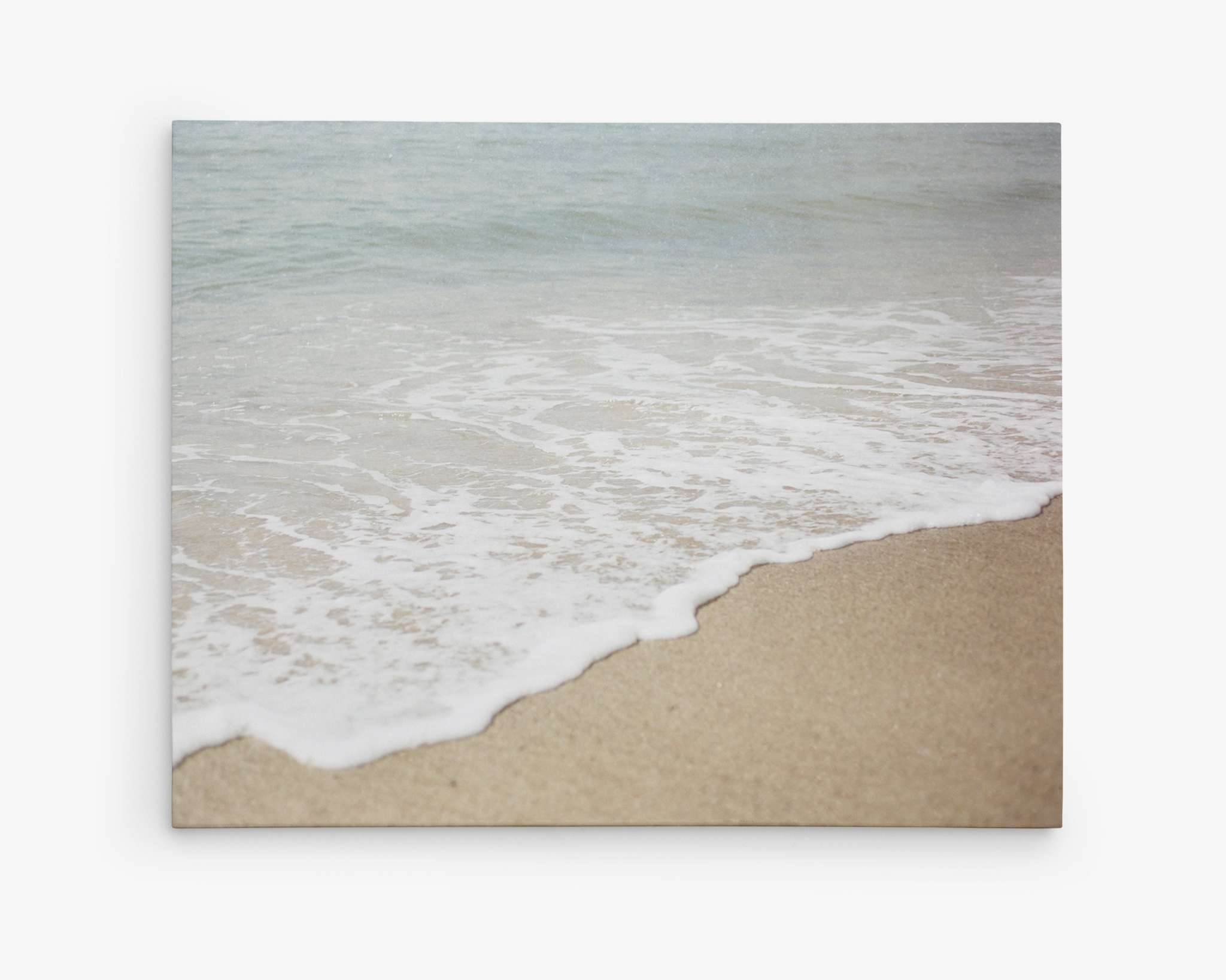 A serene beach scene of Will Rogers State Beach shows gentle waves lapping onto a sandy shore. The foam from the waves creates a white, frothy edge as it meets the beige sand, capturing a peaceful, coastal atmosphere. This image is available as Offley Green&#39;s premium artist-grade Beach Waves Canvas Wall Art, &#39;Chasing Surf,&#39; with a gallery wrap finish.
