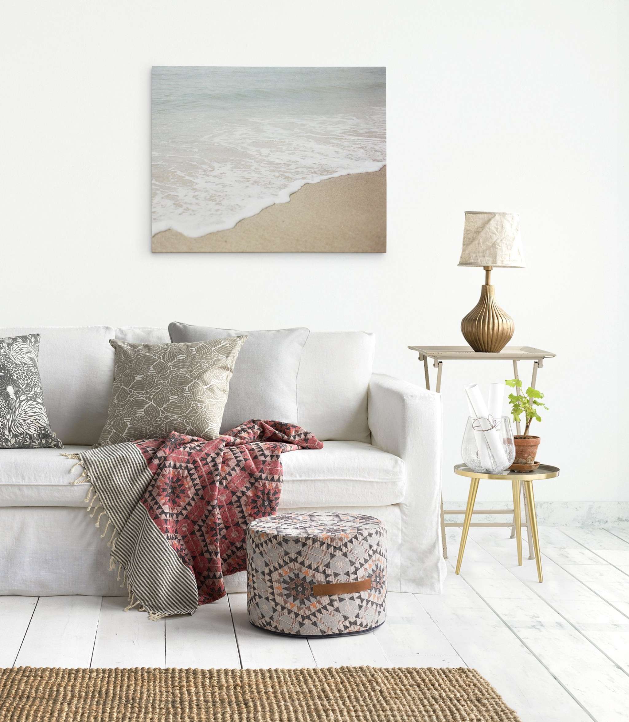 A bright living room features a white sofa with patterned cushions and a colorful throw. In front sits a patterned pouf. A side table holds a decorative lamp, a glass vase, and a potted plant. Above the sofa, an Offley Green Beach Waves Canvas Wall Art, &#39;Chasing Surf&#39; adorns the wall.