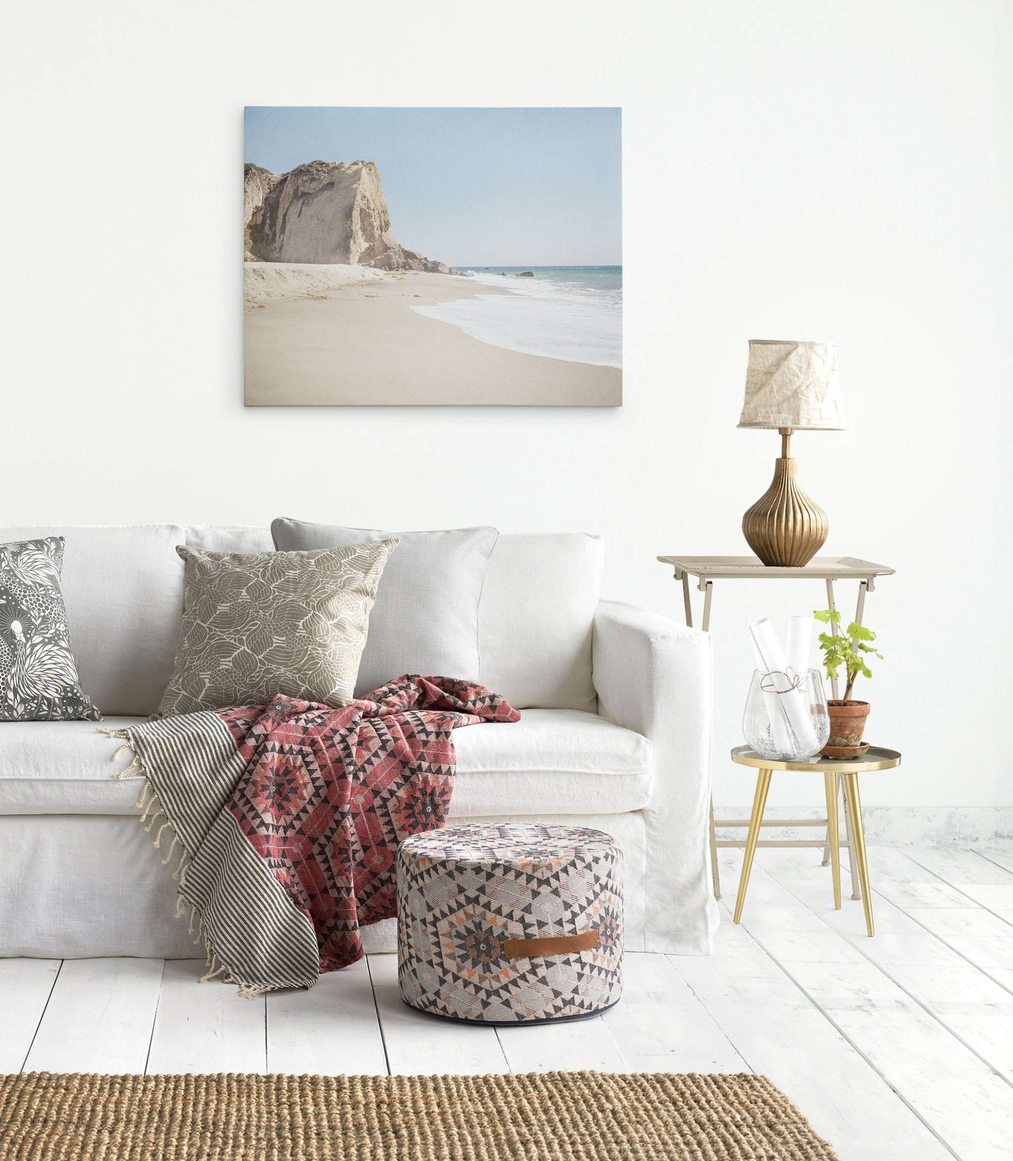 A cozy, light-filled living room features a white sofa adorned with patterned cushions and a red, geometric throw. An Offley Green California Malibu Canvas Wall Art, 'Point Dume' hangs on the white wall above. A small side table with a plant and vase, and a patterned pouf complete the decor.