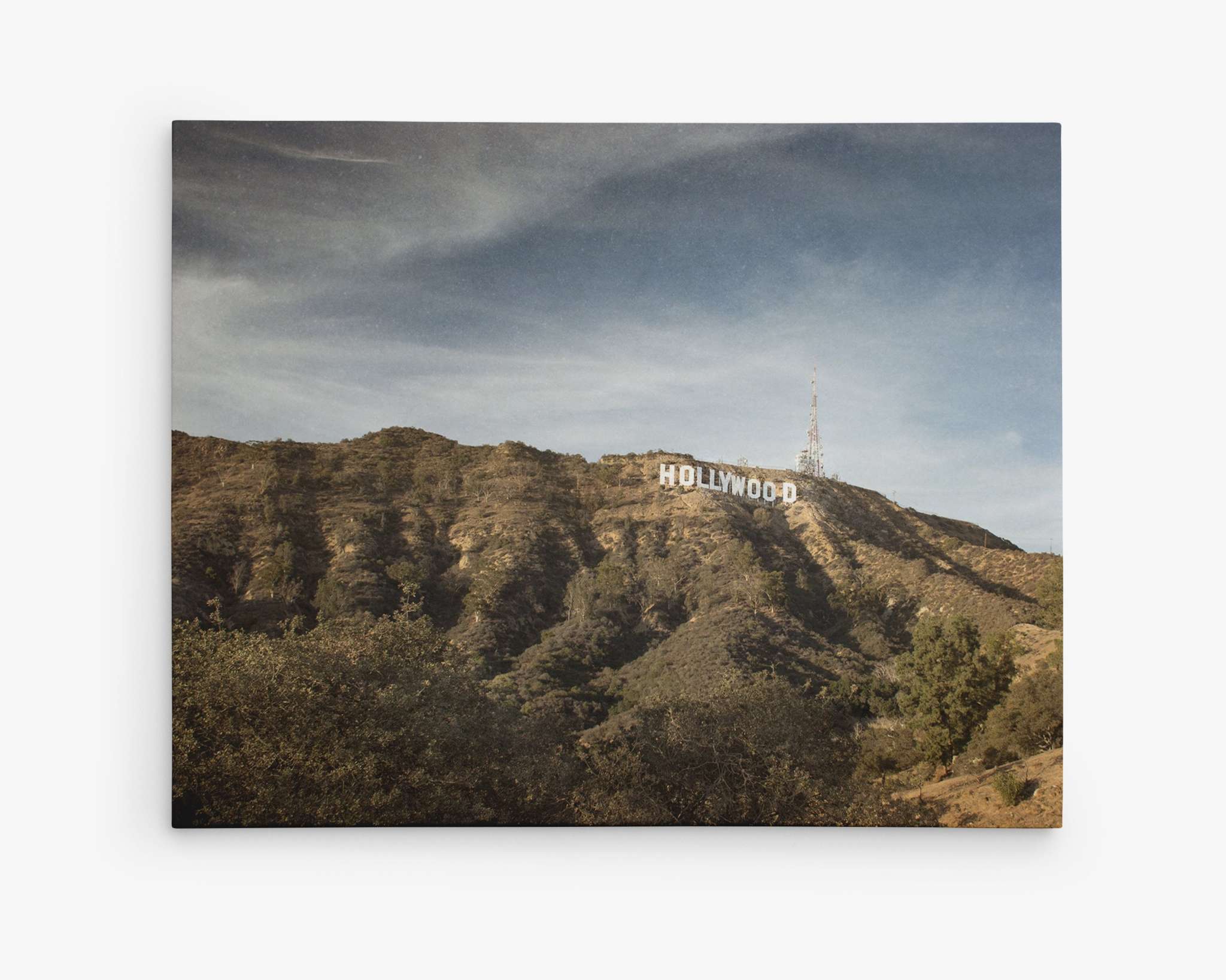 The image, showcased on a premium artist-grade canvas, depicts the iconic Hollywood sign situated on the hills with a background of a partially cloudy sky. The foreground features a mix of trees and shrubbery, highlighting the natural landscape surrounding the landmark. This stunning piece is titled **Hollywood Sign Canvas Wall Art, 'Hollywood'** by **Offley Green**.