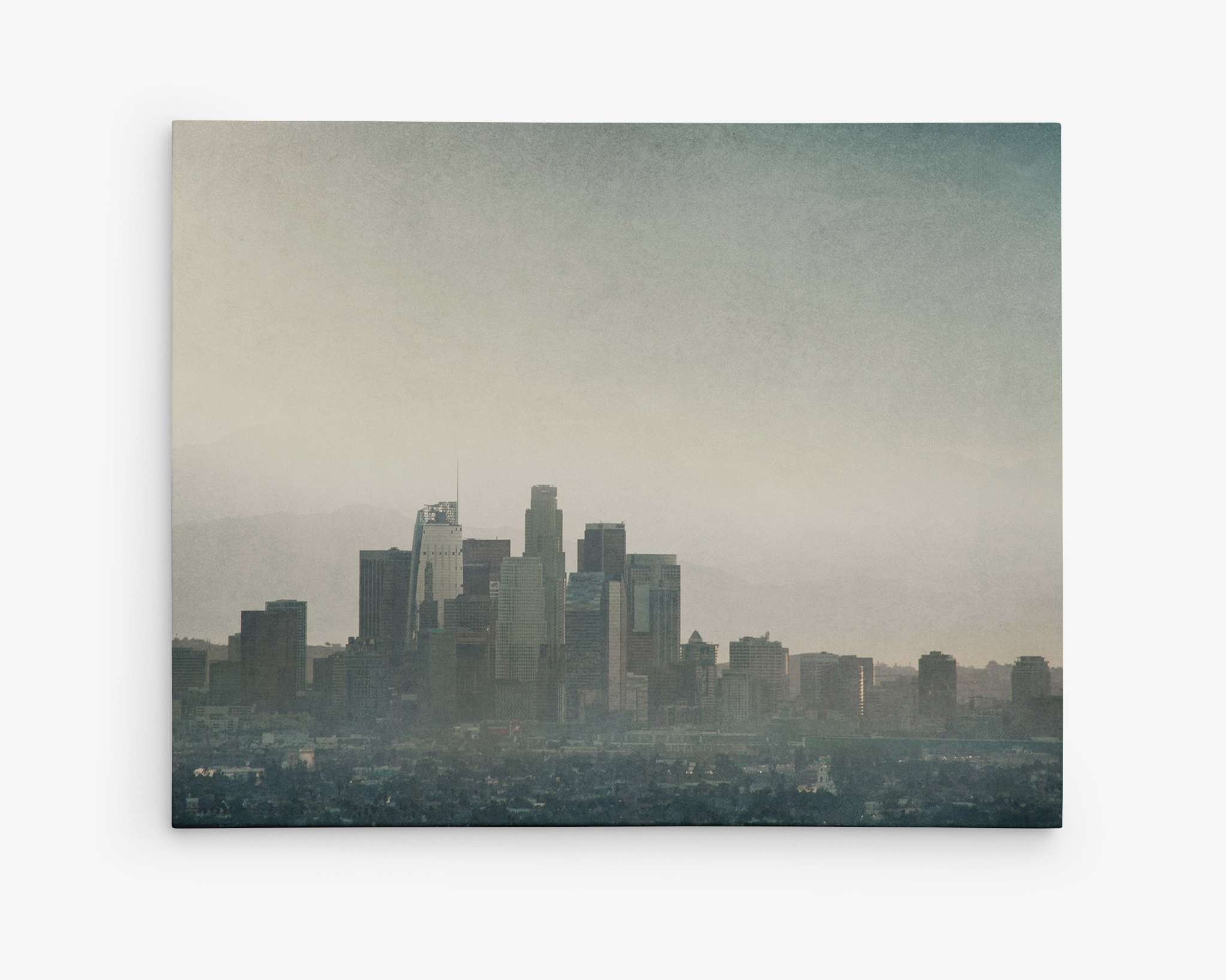 A moody downtown Los Angeles scene unfolds under a slightly hazy sky, with tall skyscrapers dominating the landscape in various heights and designs. Faintly visible through the haze is a mountainous silhouette, captured perfectly on the Downtown Los Angeles Canvas Wall Art, 'Stormy La La Land' by Offley Green for exceptional detail.