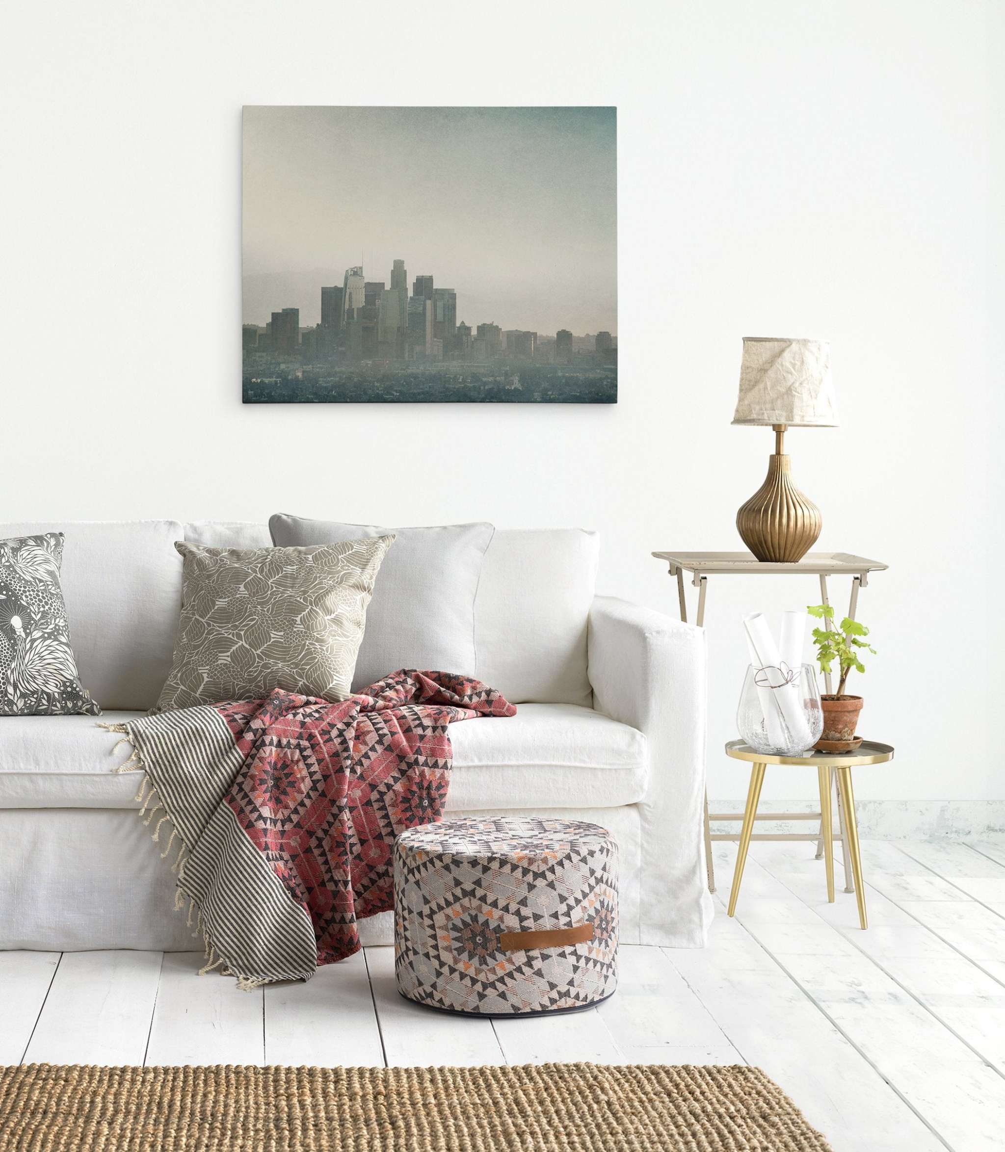 A cozy living room features a white sofa adorned with patterned cushions and a red geometric throw. A patterned pouf sits in front of the sofa. A small side table holding a lamp and a plant is placed next to it. Hanging on the wall is 'Stormy La La Land' Downtown Los Angeles Canvas Wall Art by Offley Green.