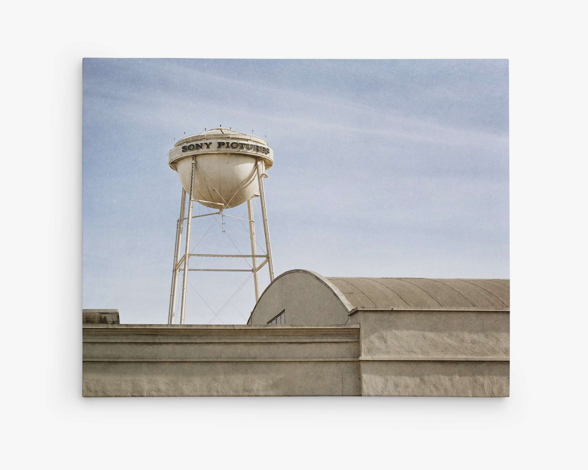 A tall water tower labeled &quot;SONY PICTURES&quot; rises above the surrounding buildings. Situated in an industrial area with lightly clouded skies, it resembles a Los Angeles Sony Pictures Studio Wall Art, &#39;Sony Lot&#39; by Offley Green. The light gray, industrial-looking buildings in the foreground complete this cinematic view of Sony Studios.