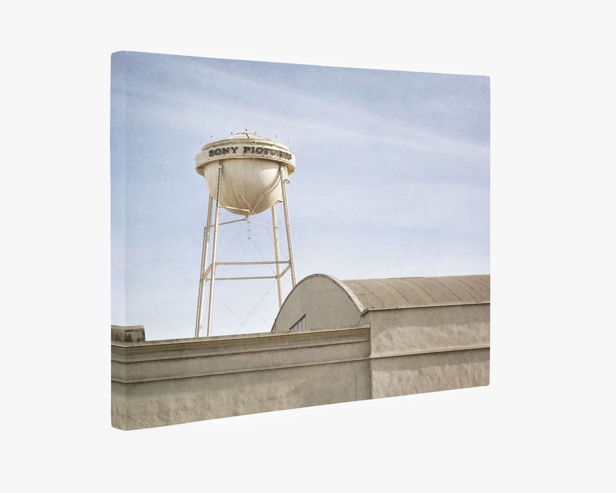 A rooftop view of a tall, white water tower with the words &quot;Sony Pictures&quot; written on it, situated against a clear blue sky. The tower stands behind a beige building with an arched roof, captured perfectly as if on an Offley Green Los Angeles Sony Pictures Studio Wall Art, &#39;Sony Lot&#39;.