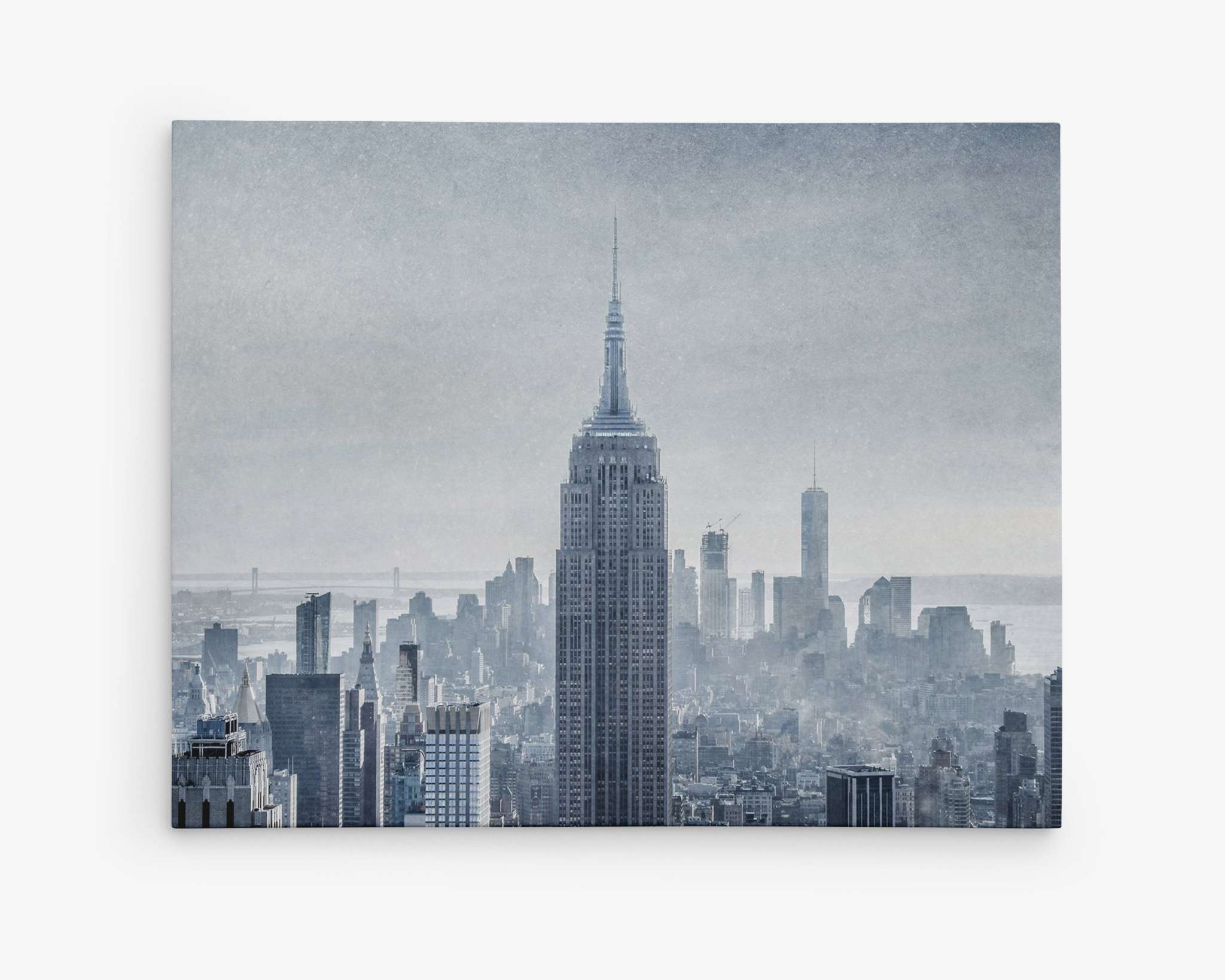 A hazy view of a city skyline dominated by the tall and iconic Empire State Building in the center is captured on the Offley Green New York City Canvas Wall Art, 'Winter Metropolis'. The surrounding buildings appear under a soft, foggy atmosphere, blending into the misty background, giving a sense of depth and scale to the urban landscape.
