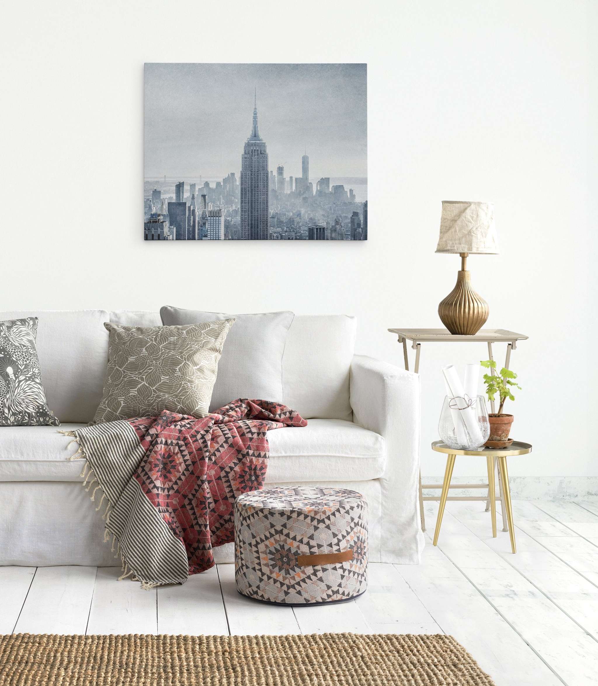 A cozy living room with a white sofa adorned with patterned cushions and a red-patterned throw blanket made of premium poly-cotton blend. A round patterned ottoman sits nearby. A small side table with a lamp, plant, and books is on the right, and an Offley Green New York City Canvas Wall Art titled 'Winter Metropolis' hangs on the white wall behind the sofa.