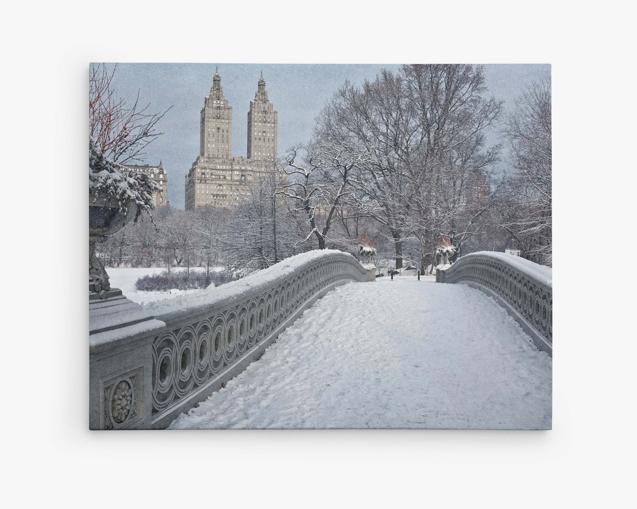 A snow-covered Bow Bridge leads into Central Park in New York City during winter. The trees are dusted with snow, and two iconic tall buildings are visible in the background, standing against a gray sky. The scene is quiet, serene, and picturesque—perfect for the Offley Green New York City Canvas Wall Art, 'Snow on Bow Bridge'.