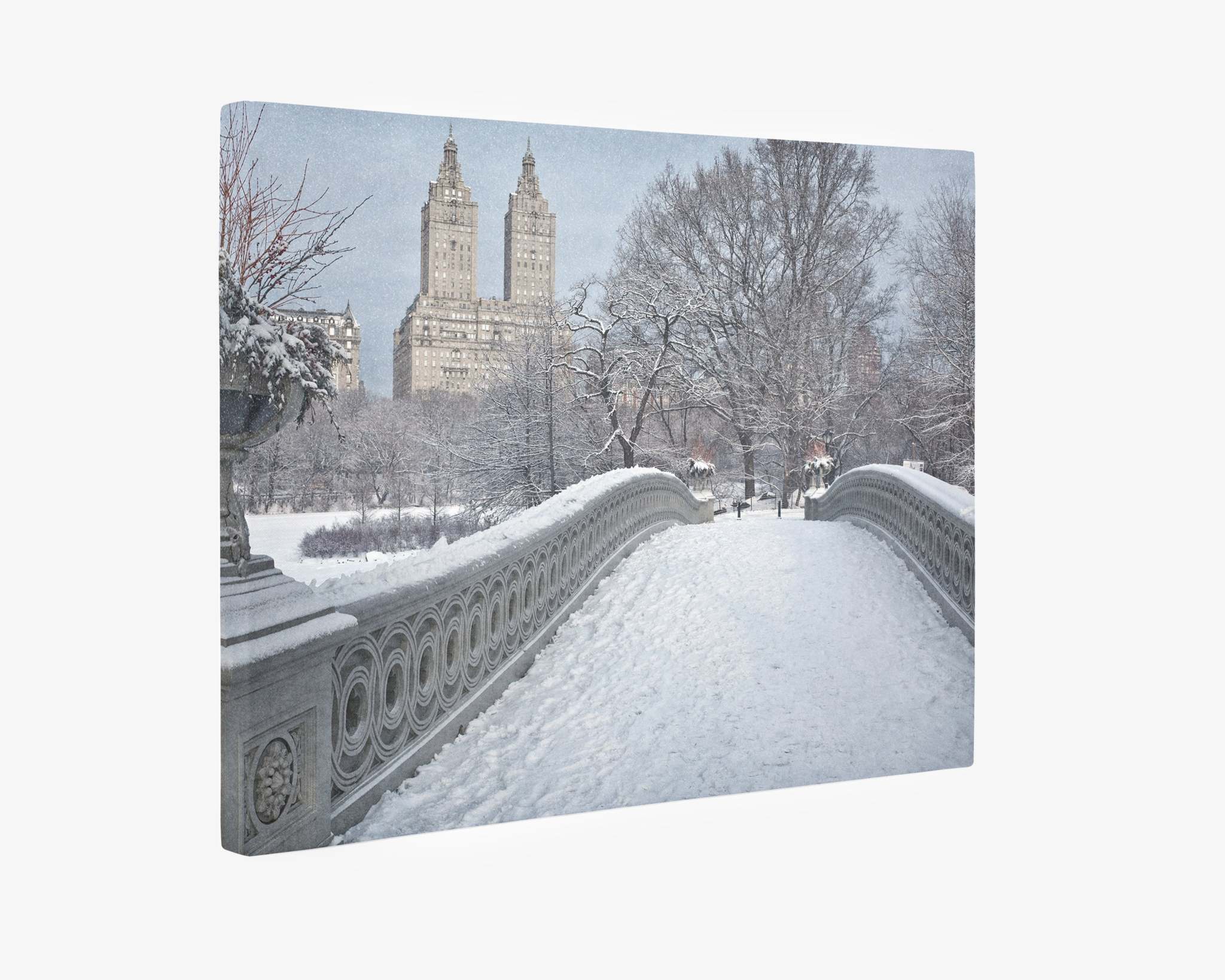 A snow-covered Bow Bridge in Central Park, New York City, leads the viewer's eye towards two prominent, twin-towered buildings in the background. Trees laden with snow frame the scene, adding to the wintery atmosphere. The sky is overcast, enhancing the serene ambiance perfect for a New York City Canvas Wall Art, 'Snow on Bow Bridge' by Offley Green.