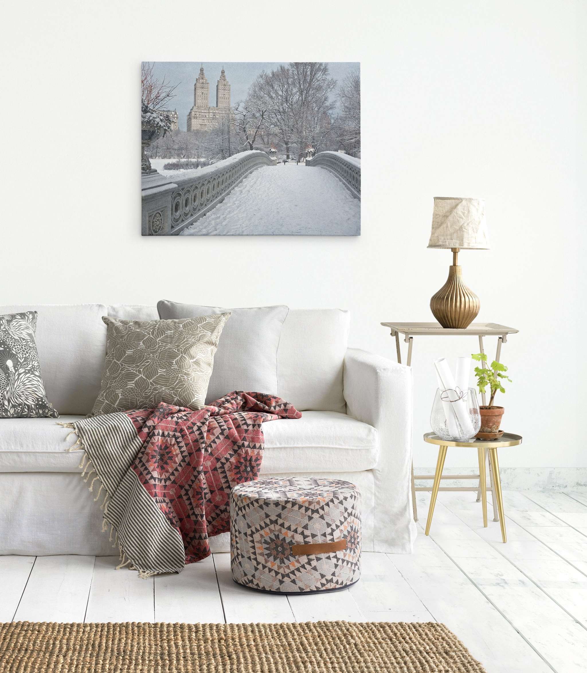 A cozy living room features a light beige sofa adorned with patterned pillows and a red and gray blanket. A patterned ottoman and rug complement the space. Beside the sofa is a small table with a lamp, plant, and books. An Offley Green New York City Canvas Wall Art, 'Snow on Bow Bridge' hangs on the white wall.