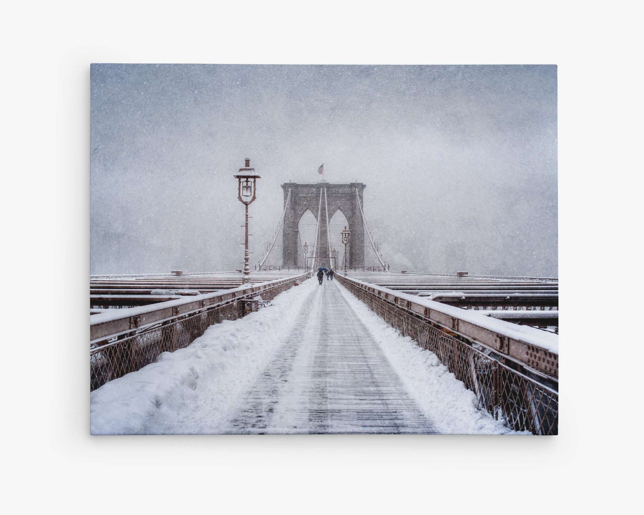 A snow-covered Brooklyn Bridge in New York City during heavy snowfall resembles a scene straight out of a Christmas card. The walkway is lined with snow, and a few people are seen in the distance walking towards the iconic twin arches, all beneath an overcast sky that creates a serene, wintery atmosphere. This picturesque view perfectly captures the essence of Offley Green's New York Brooklyn Wall Art, 'Brooklyn Snow.'