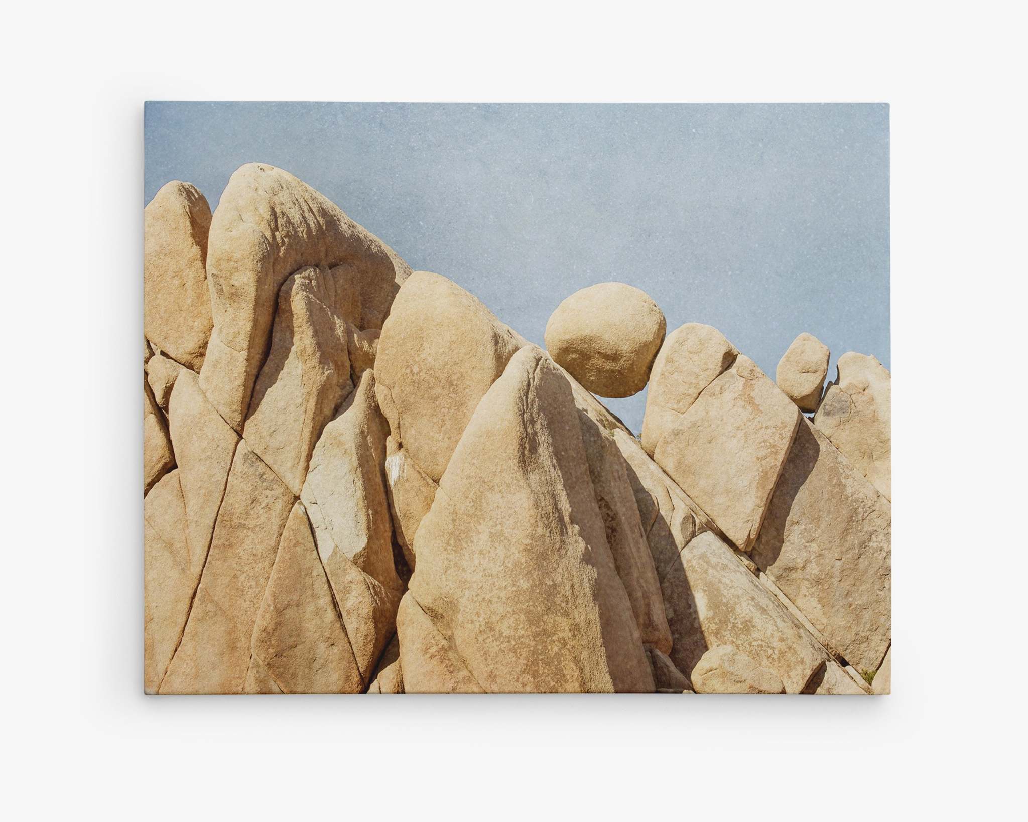 A collection of large, smooth, tan Joshua Tree desert rock formations against a clear blue sky. One round boulder appears to balance delicately atop a pointed rock, creating a captivating and precarious scene in a desert landscape—a perfect subject for Offley Green's Joshua Tree Canvas Wall Art, 'Rock Formations'.