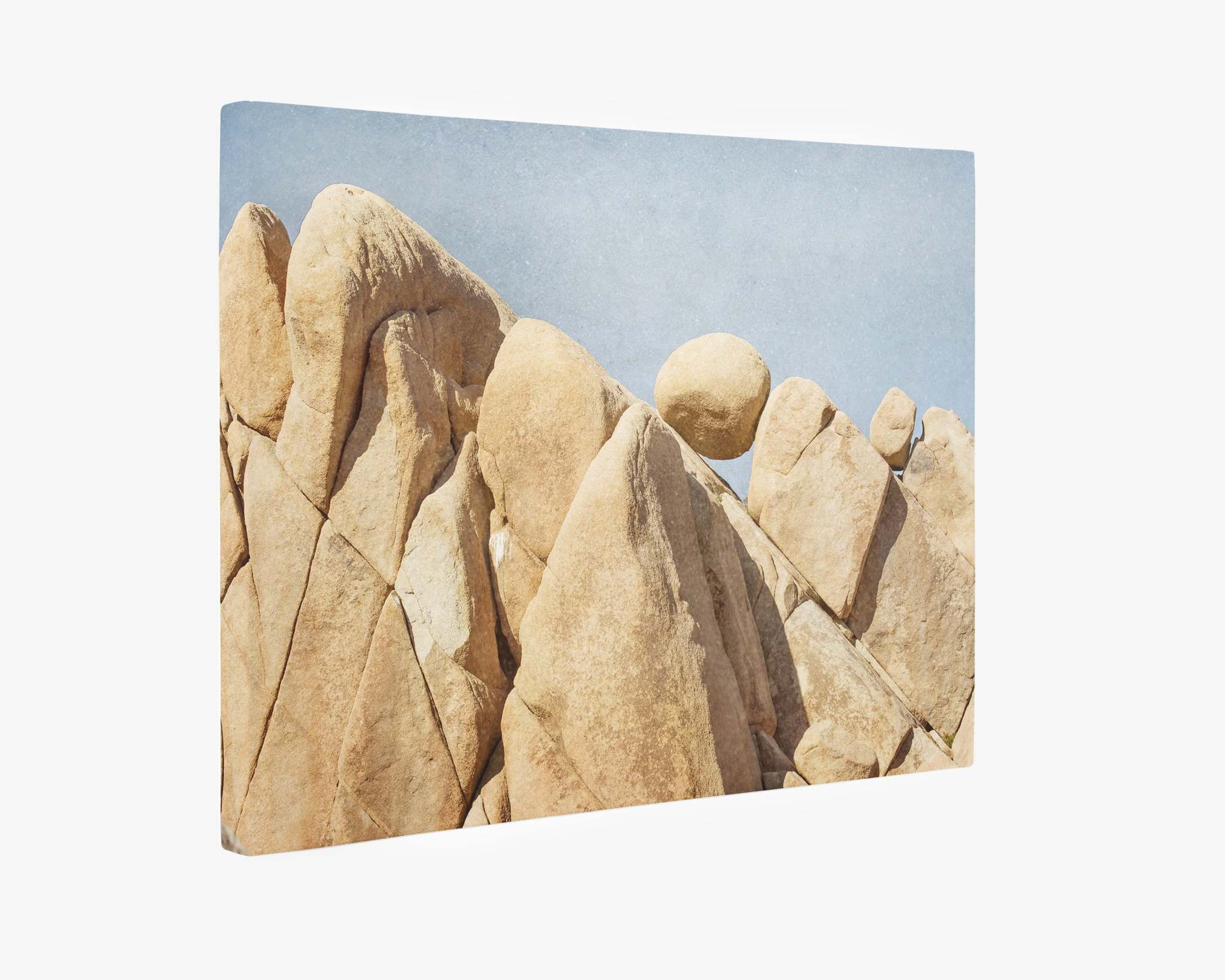 A rugged landscape of large, tan Joshua Tree desert rock formations under a clear blue sky. One round boulder appears to be precariously balanced atop a pointed rock. The natural scene conveys a sense of tranquility and geological wonder, perfect for Offley Green's Joshua Tree Canvas Wall Art, 'Rock Formations,' reflecting nature's beauty.