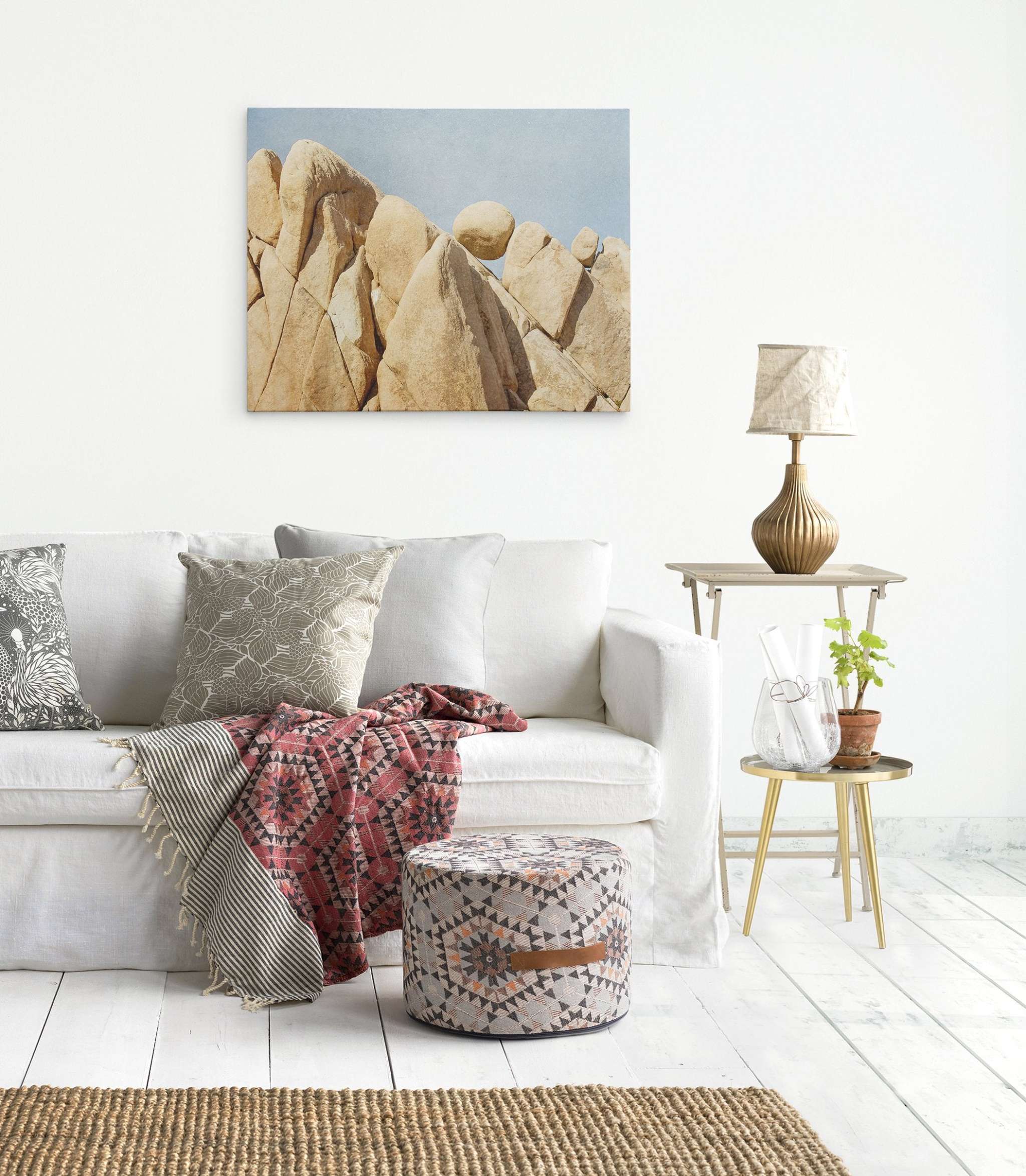 A minimalist living room features a white sofa adorned with patterned pillows and a red geometric throw blanket. A patterned ottoman and a small side table with a lamp, potted plant, and books sit nearby. A Joshua Tree Canvas Wall Art, 'Rock Formations' by Offley Green hangs on the white wall.