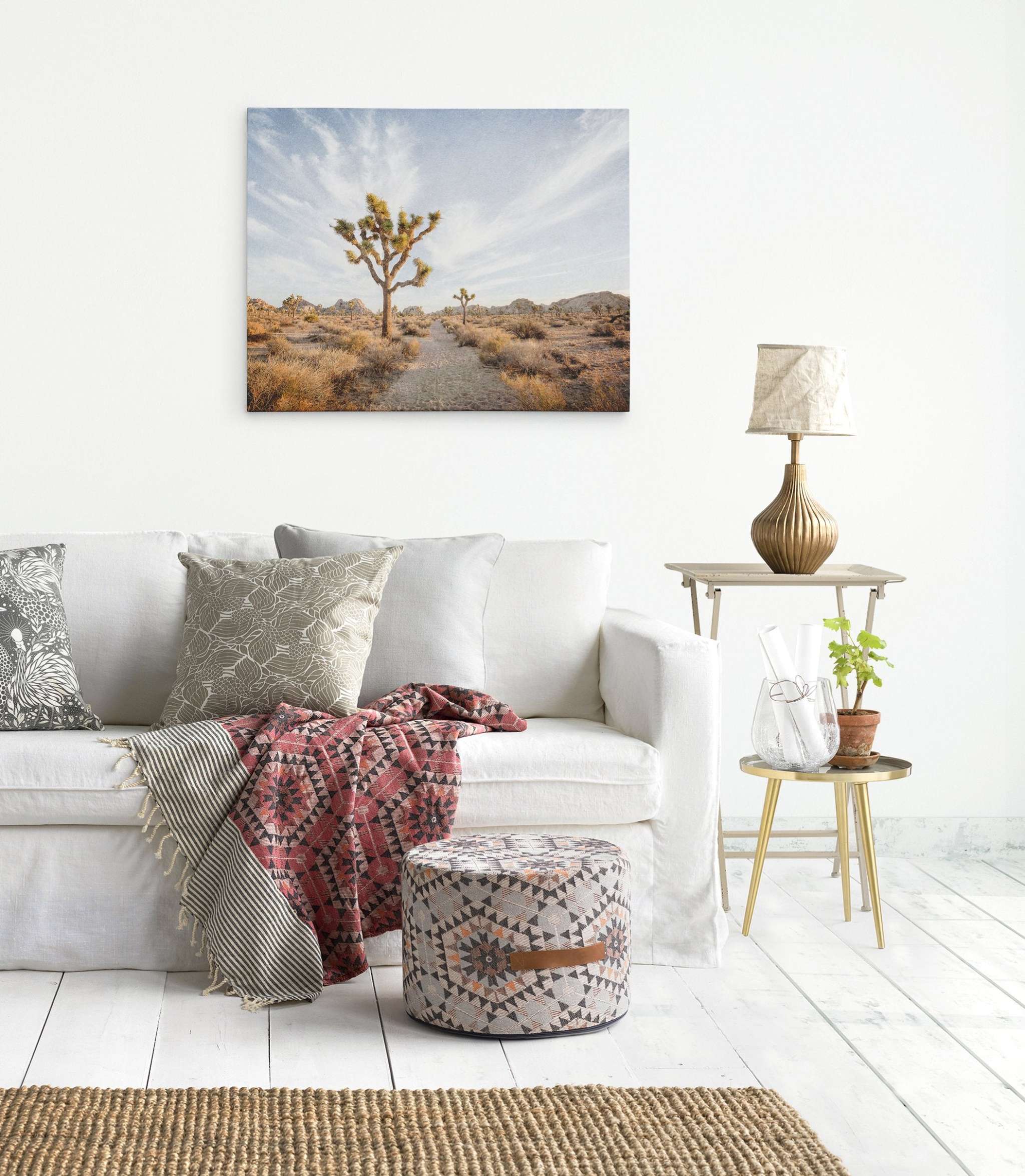 A cozy living room features a white sofa adorned with various patterned cushions and a textured throw. A round pouf with geometric designs sits in front. A side table holds a lamp, potted plant, and glass vase. Above the sofa, an Offley Green Joshua Tree Canvas Wall Art, 'Path to Joshua' completes the setting.