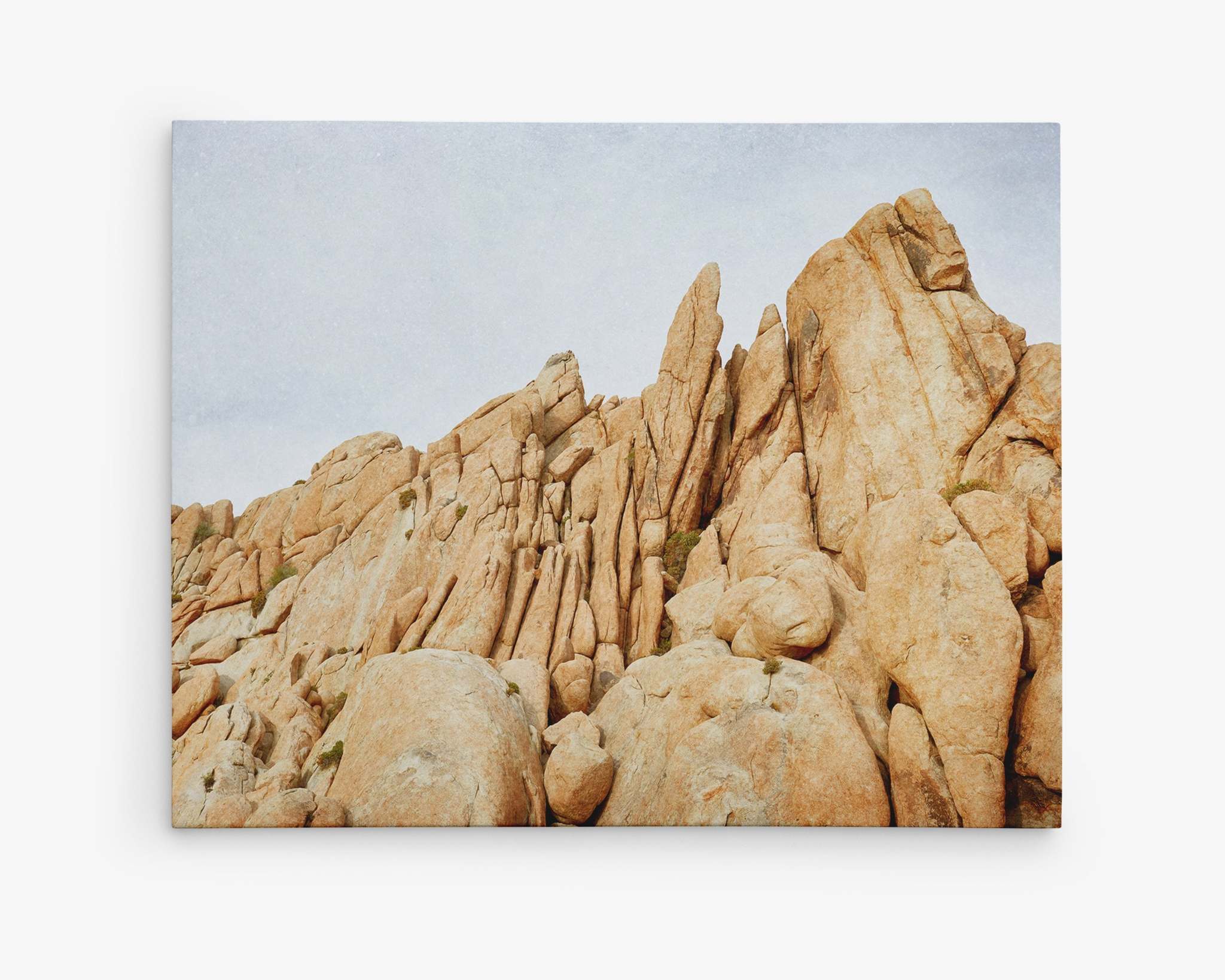 A rugged, rocky landscape featuring large, jagged sandstone formations under a pale, cloudy sky. The orange-hued rocks are stacked unevenly, resembling mid-century Palm Springs architecture, creating a striking natural scene perfect for Offley Green's Joshua Tree Canvas Wall Art titled 'Joshua Rocks'.